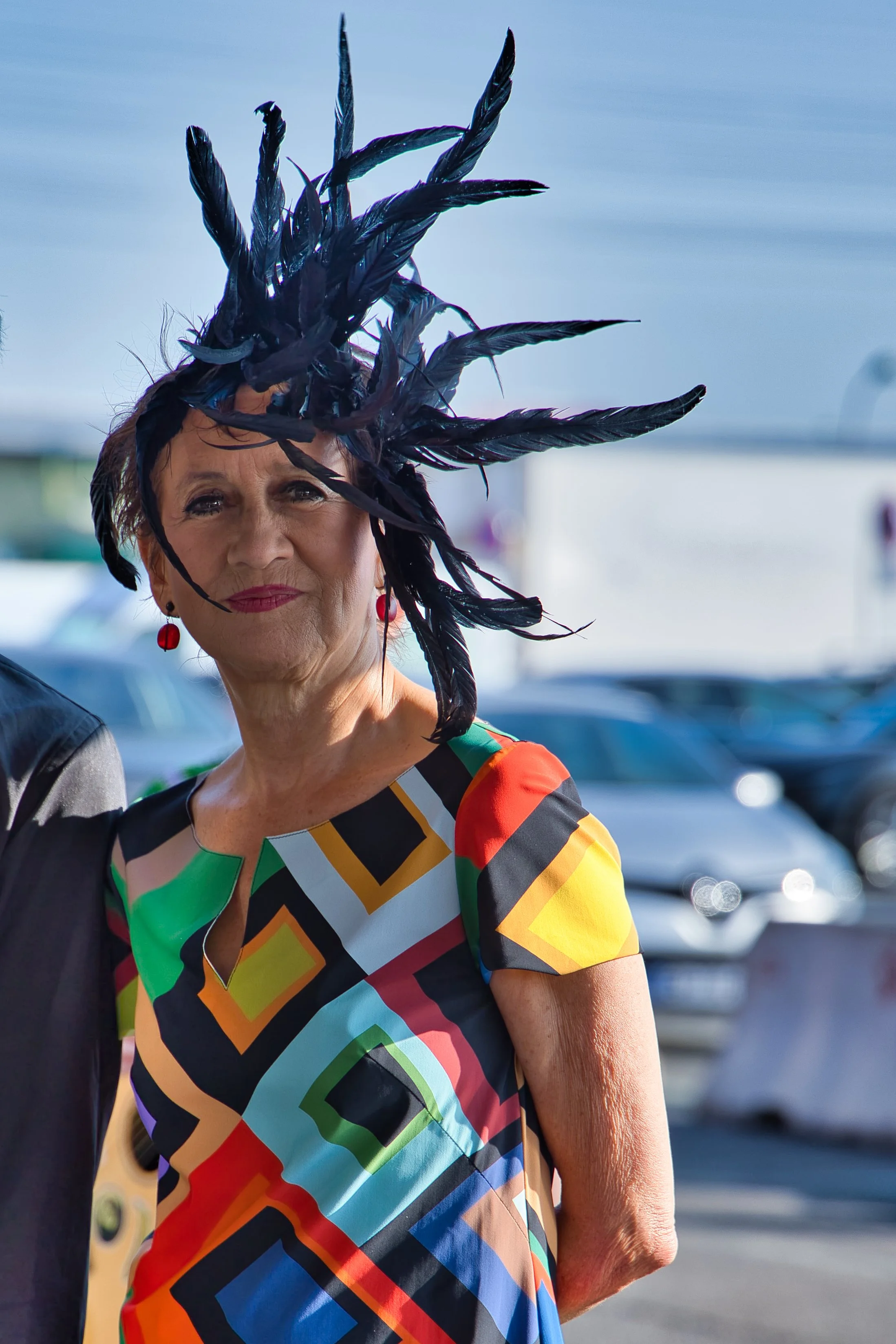 Une femme portant un chapeau noir avec des plumes et une robe à motifs géométriques colorés, posant lors d'un événement en plein air.