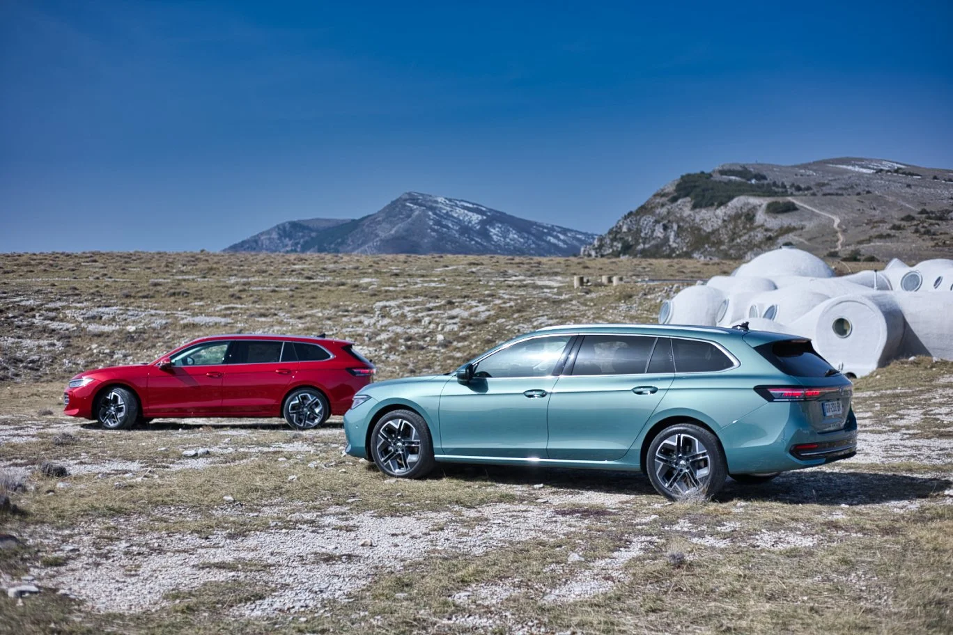 Volkswagen Passat rouge et bleu-vert, stationnées sur un terrain rocailleux avec des montagnes en arrière-plan et des structures architecturales modernes blanches à droite.