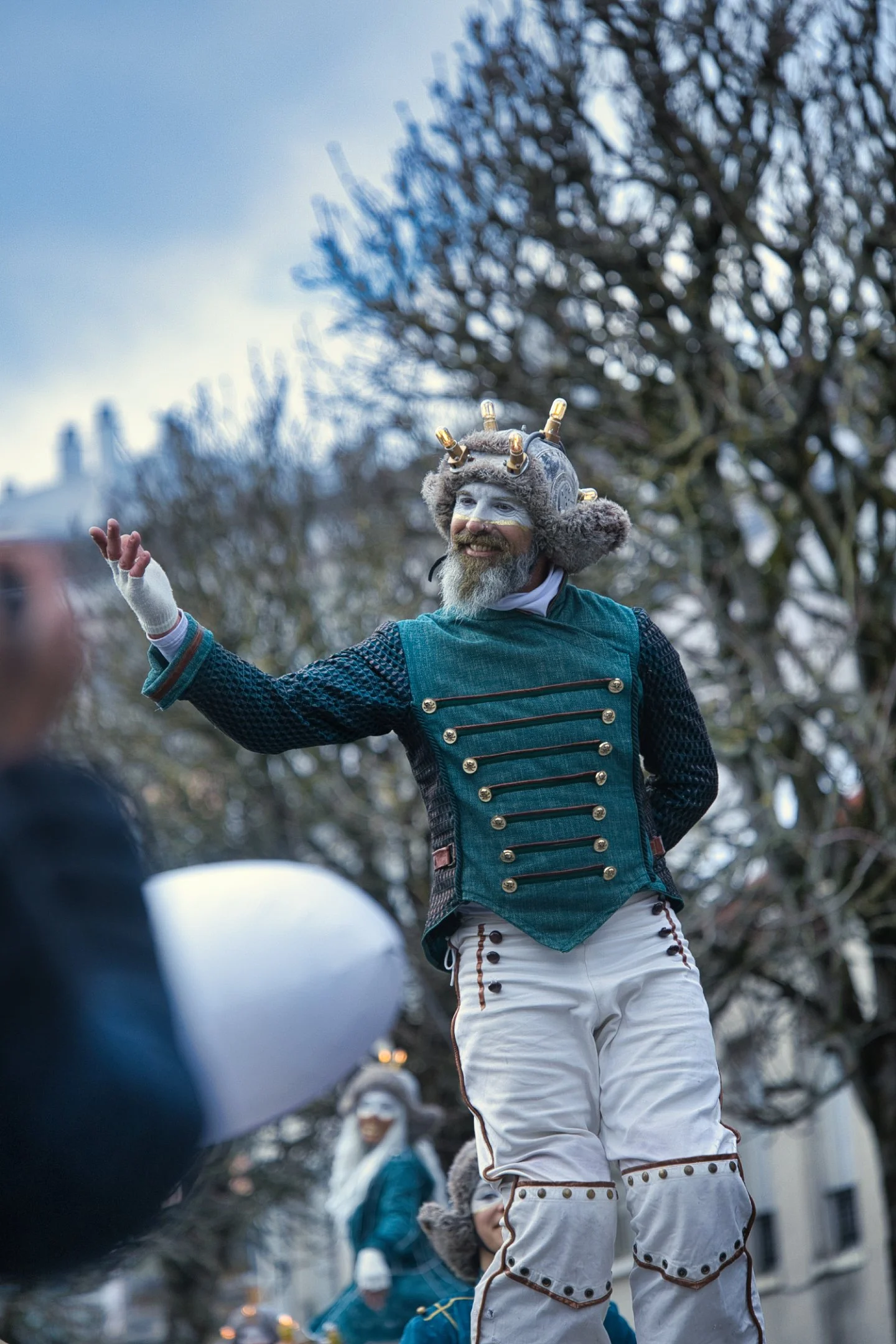 Une personne déguisée en créature fantastique avec un chapeau orné de petites ampoules, un masque sur le visage, portant un costume de style historique, participe à un carnaval dans une rue.