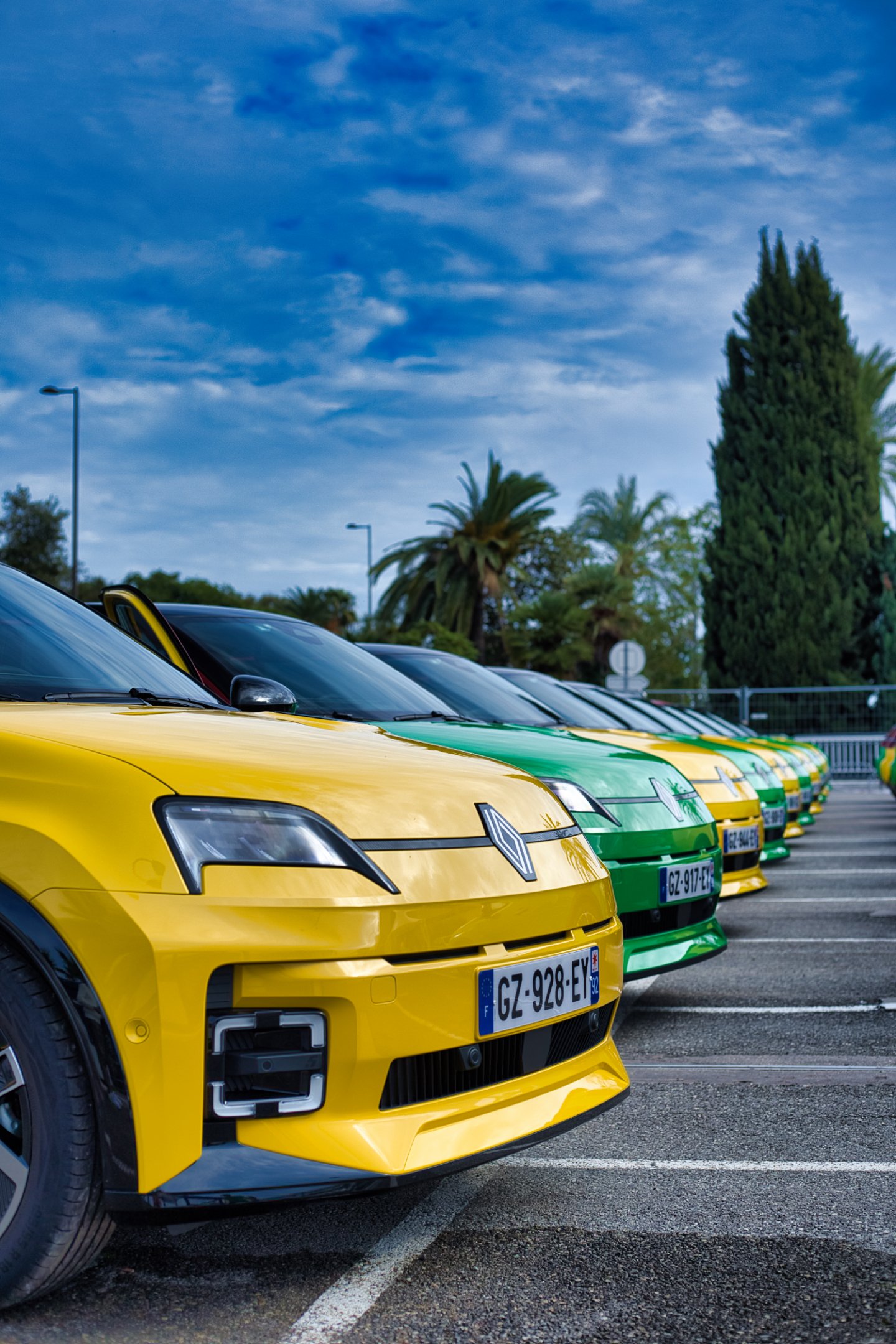 Renault R5 e-tech colorées jaunes et vertes dans un parking, avec un ciel nuageux en arrière-plan.