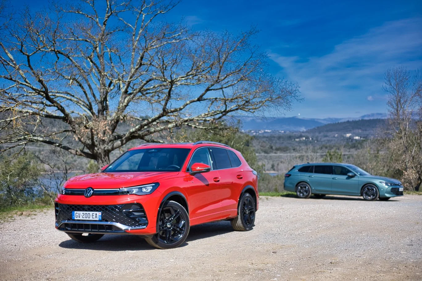 Deux voitures Volkswagen, l'une rouge au premier plan et une autre gris-vert derrière, garées sur un chemin de gravier à la campagne avec des arbres nus, une rivière, des montagnes et un ciel bleu. Volkswagen Tiguan Passat