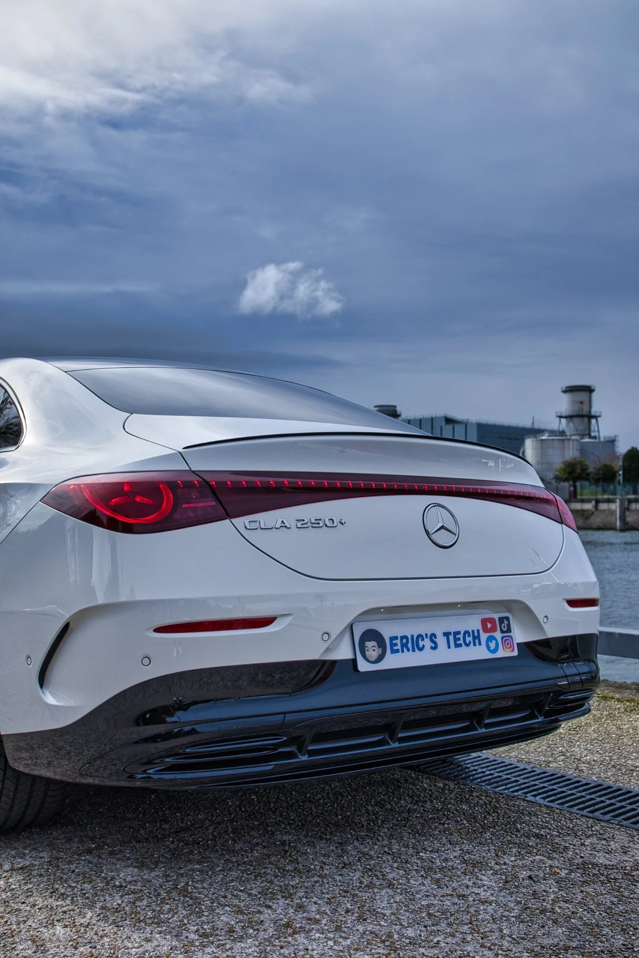 Voiture Mercedes-Benz CLA 250 sur le bord de l'eau, ciel nuageux, bâtiment industriel en arrière-plan, plaque d'immatriculation avec le texte 'ERIC'S TECH'.