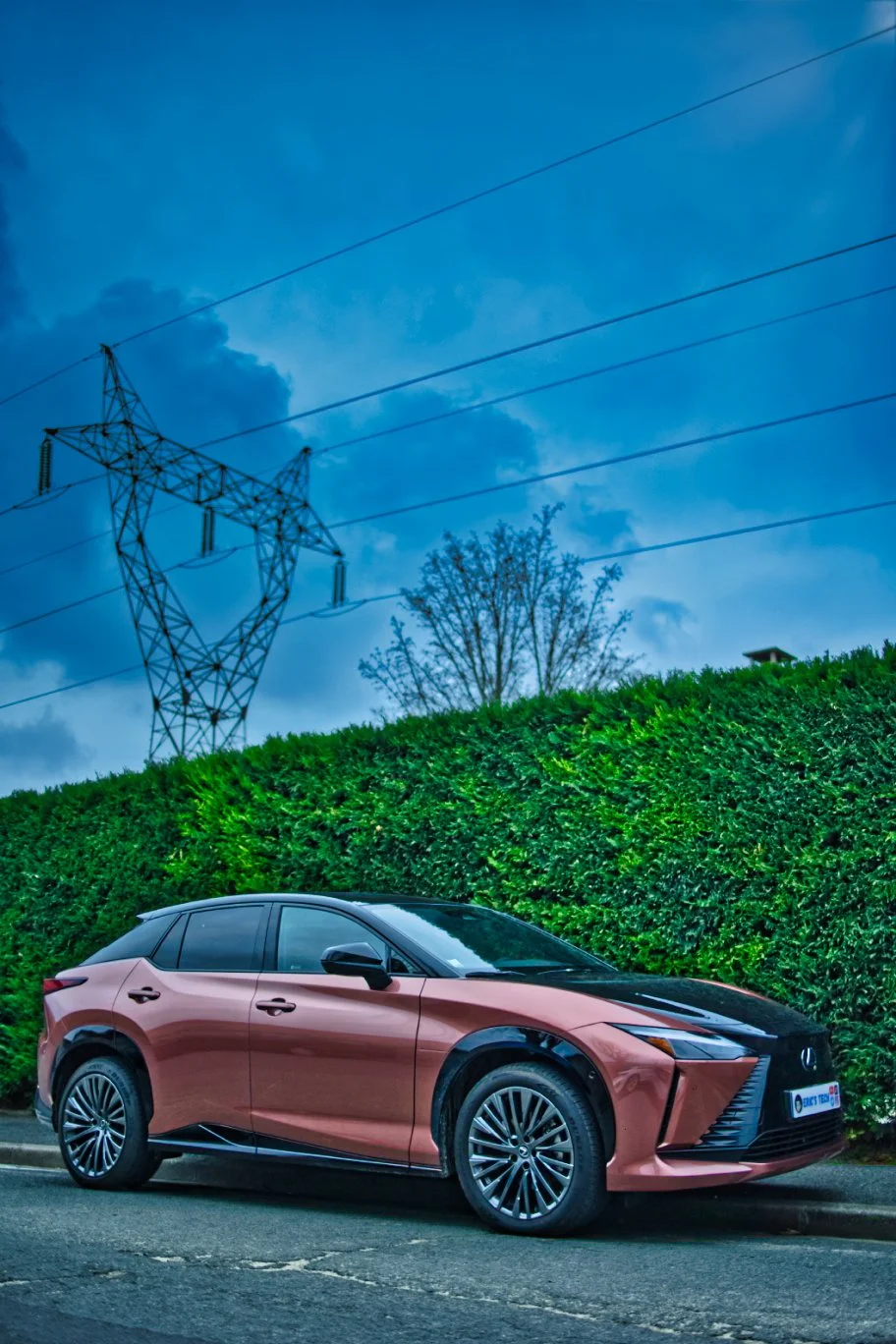 Lexus RZ 450e en stationnement sur le côté d'une route, avec un grand buisson de verdure en arrière-plan et un ciel nuageux au-dessus.