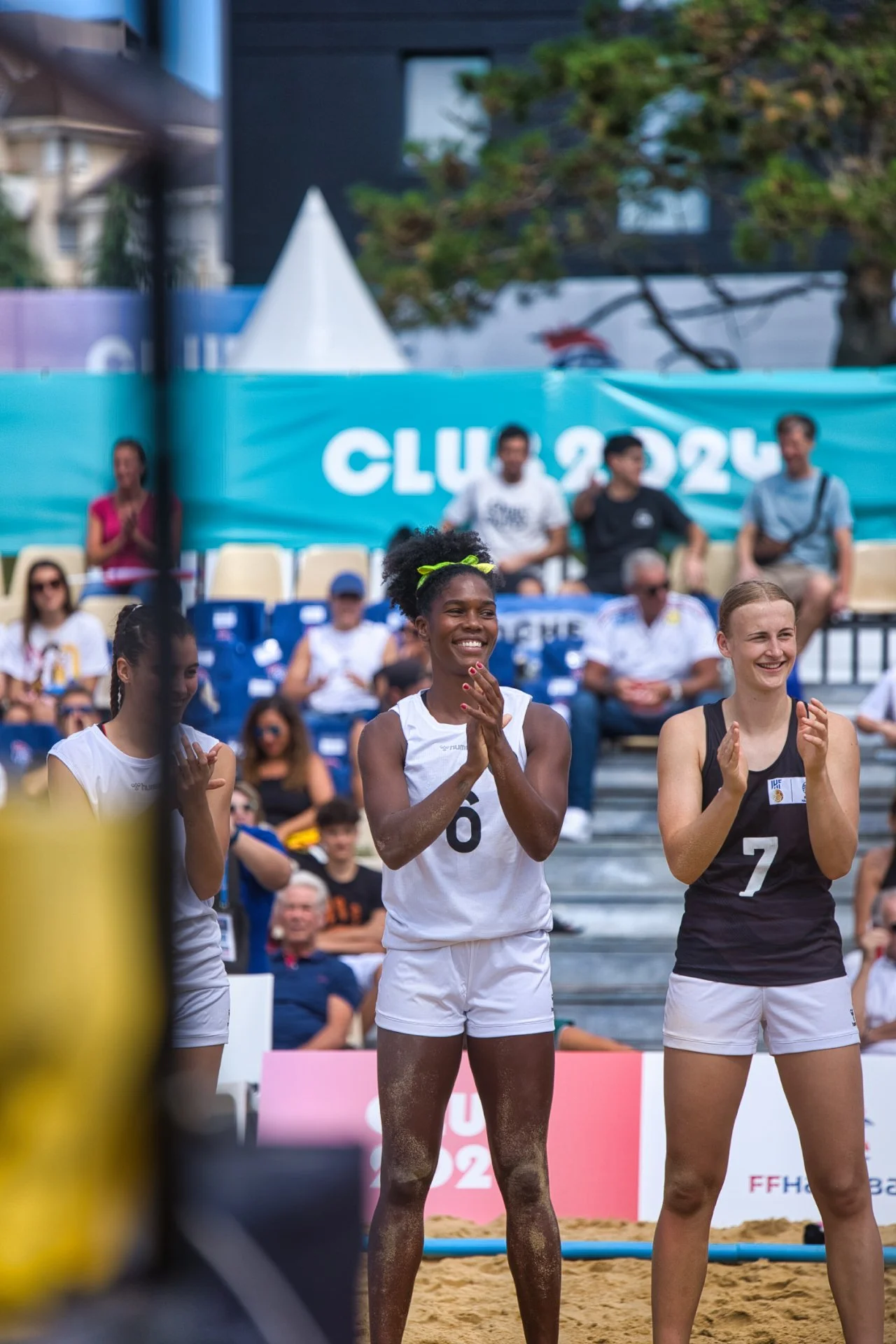 Trois joueuses de beach-volley en tenue de sport applaudissant, avec un public en arrière-plan et une bannière bleue indiquant 'CLUB 2024'.