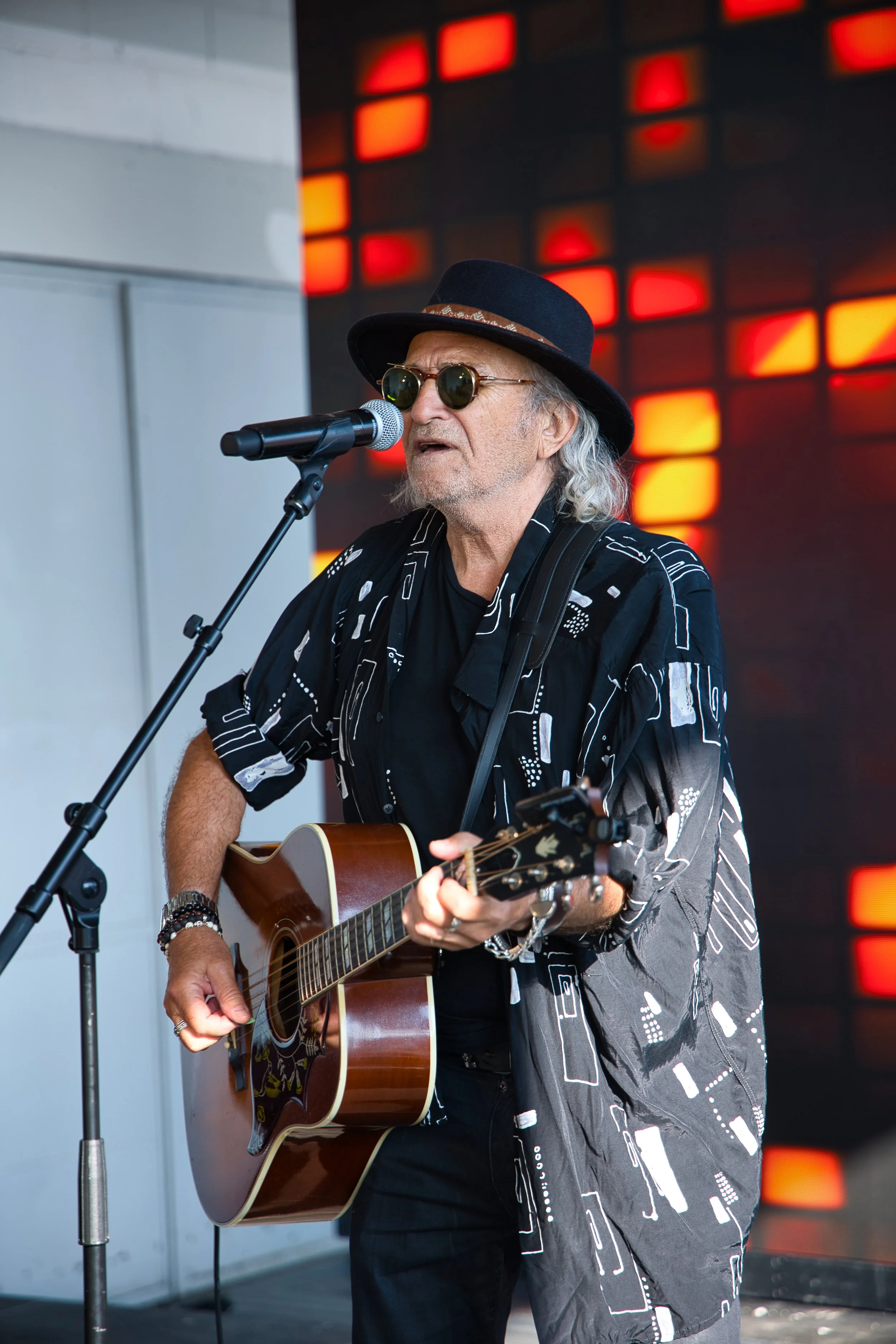 Un homme âgé avec des cheveux gris, portant un chapeau noir, des lunettes rondes et une chemise noire avec des motifs blancs, joue de la guitare acoustique et chante devant un micro, avec un fond coloré rouge et orange.