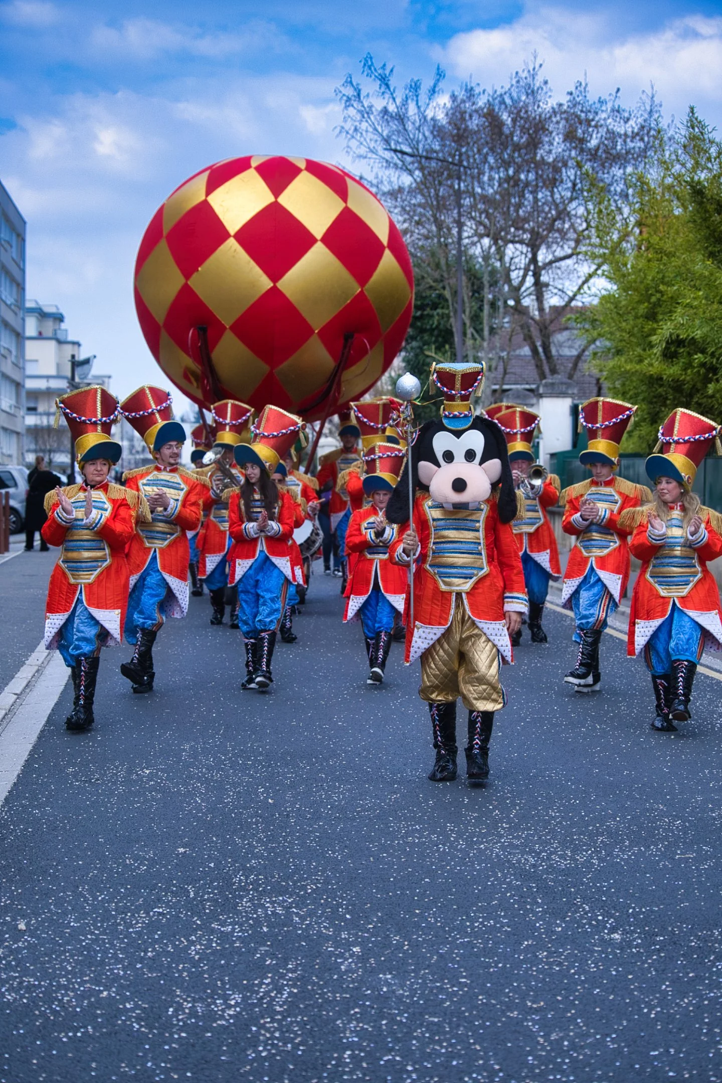 Personnage déguisé en Mickey Mouse dans un défilé avec un groupe de musiciens habillés en costumes colorés, marchant dans une rue bordée d'arbres et de bâtiments, avec un ballon géant en arrière-plan lors d'un carnaval.