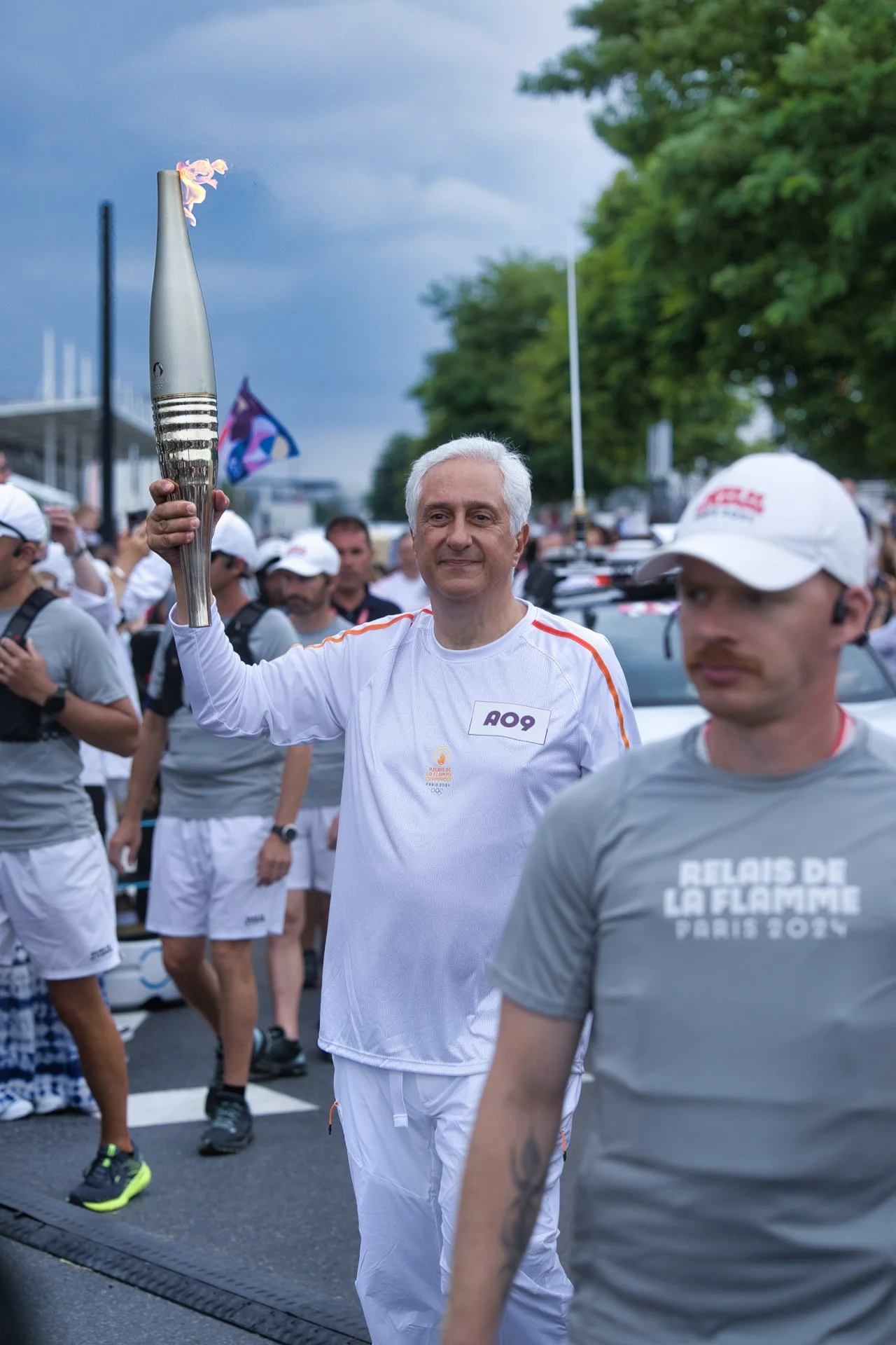 Une personne souriante tient une flamme olympique lors de la cérémonie d'ouverture d'une événement sportif, avec un groupe de participants en arrière-plan, sous un ciel nuageux. JO Paris 2024