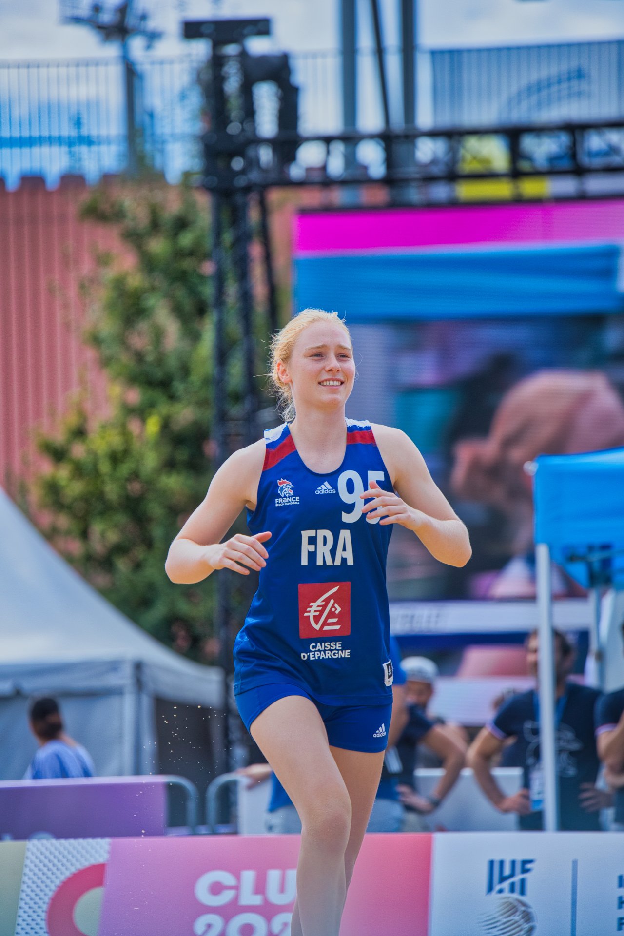 Une athlète féminine lors d'une compétition de volleyball en plein air, portant un maillot bleu avec le numéro 95 et le drapeau de la France, souriant en courant.