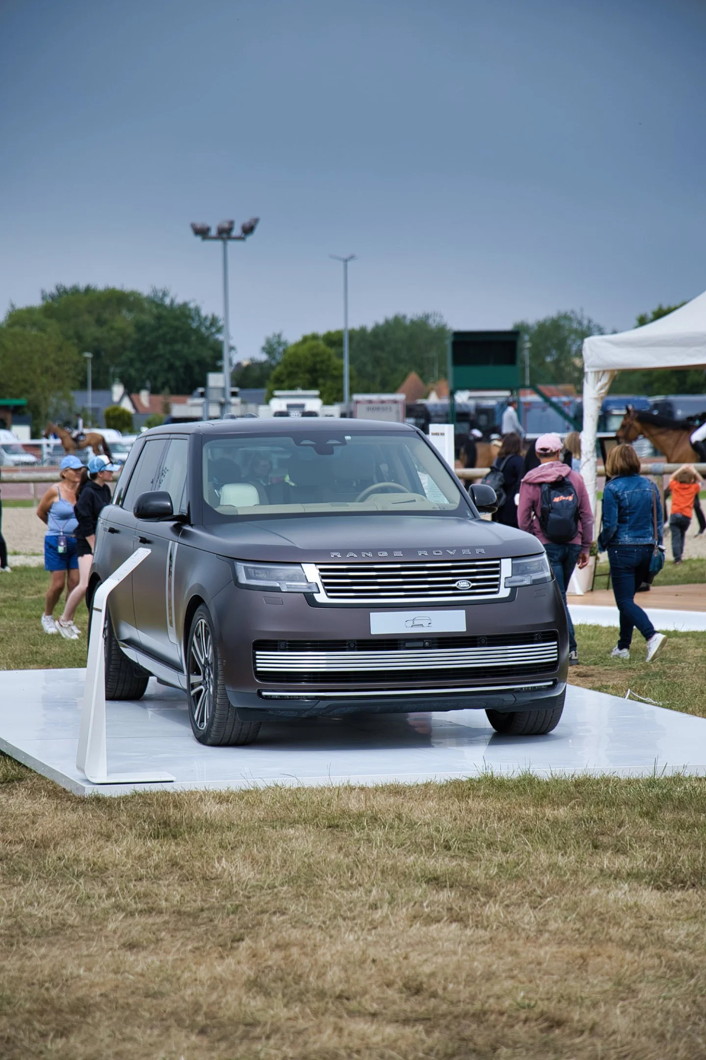 Une voiture Range Rover noire exposée lors d'un événement en plein air, avec des personnes qui varient, l'arrière-plan montre des chevaux et des stands.
