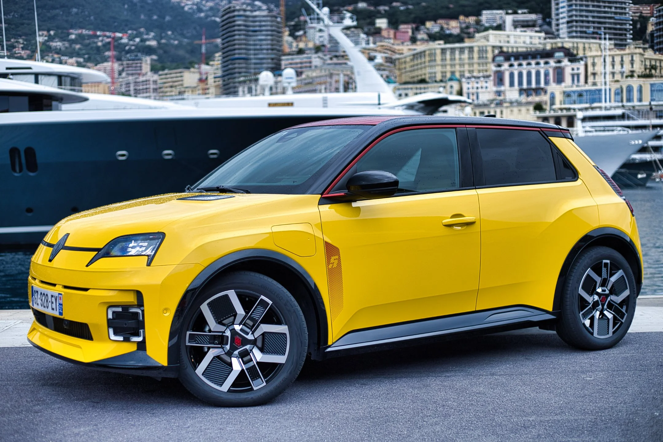 Renault R5 e-tech jaune avec des accents noirs garée près d'un port, avec un yacht en arrière-plan et une ville en collines.