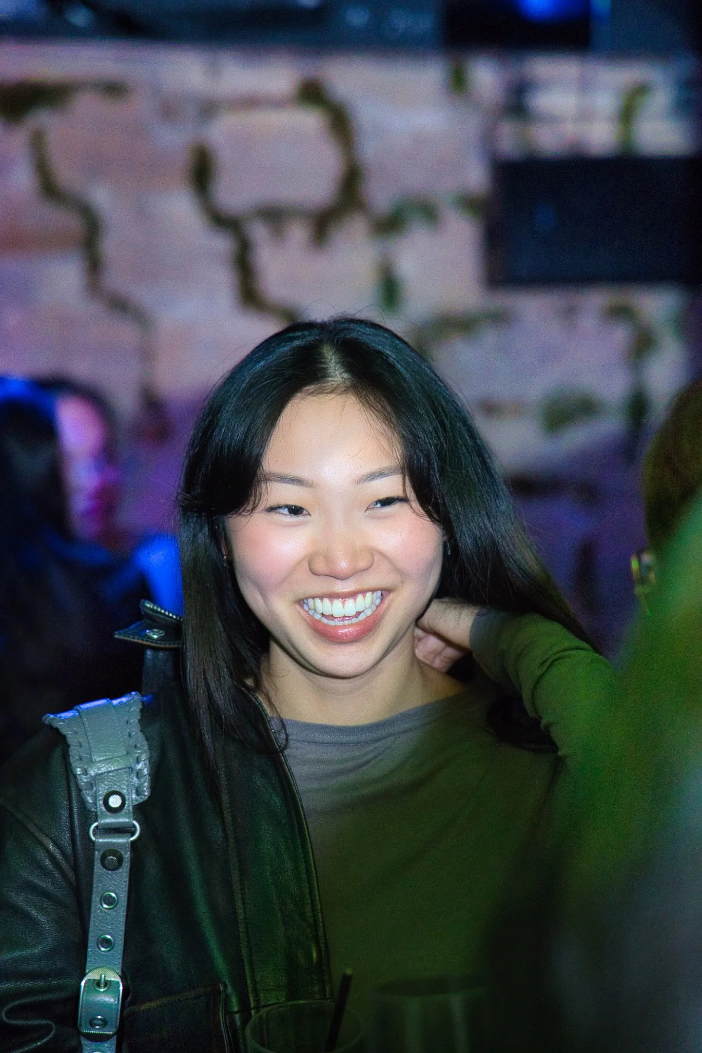 Une jeune femme souriante avec les cheveux noirs longs, portant un manteau en cuir noir et un haut gris, lors d'une sortie nocturne dans un lieu avec des murs en pierre décorés de plantes vertes et de lumières colorées.