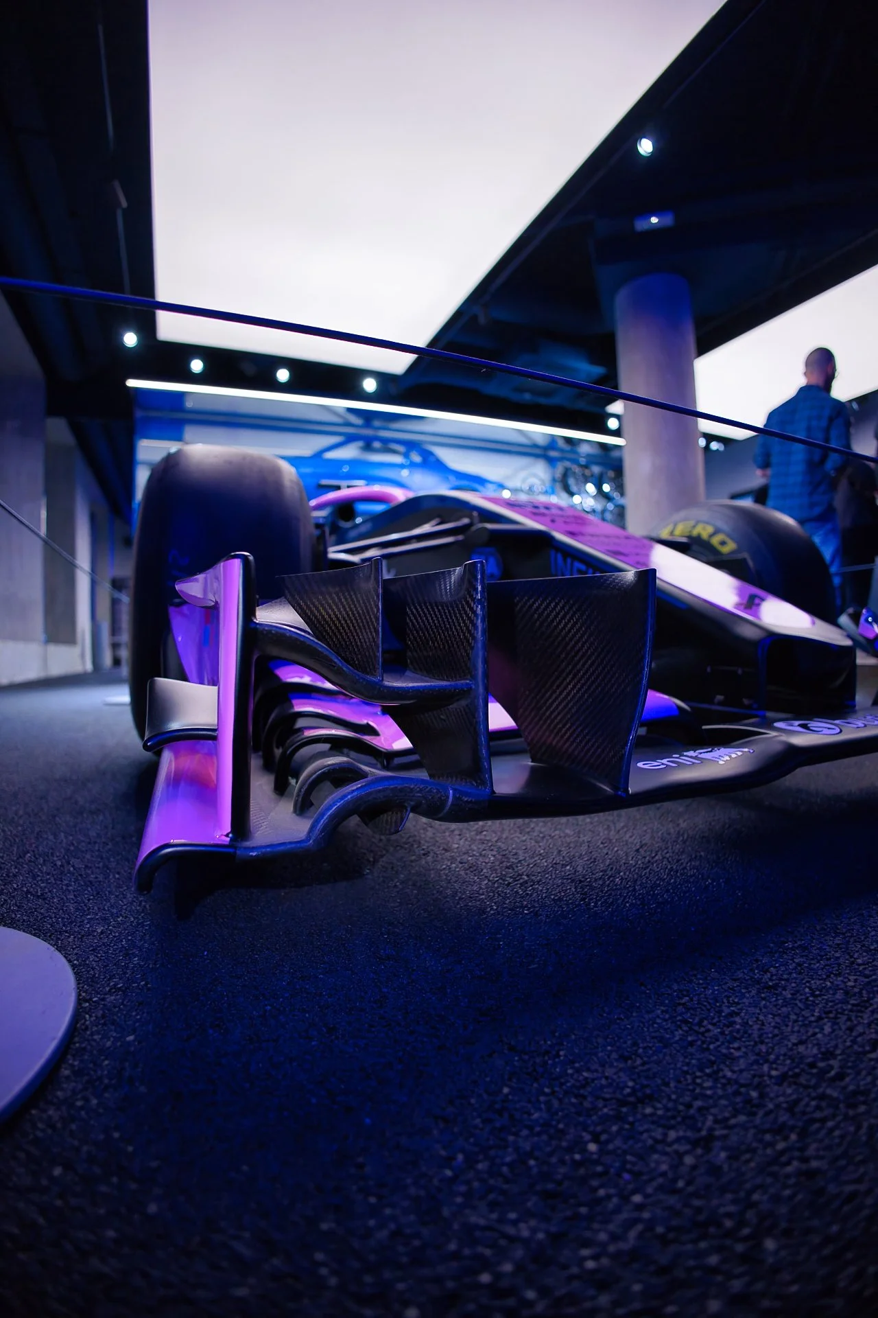 Une voiture de course de sport exposée en intérieur, vue de face, avec un focus sur la partie avant et l'aileron en carbone, éclairée par un éclairage violet et bleu.