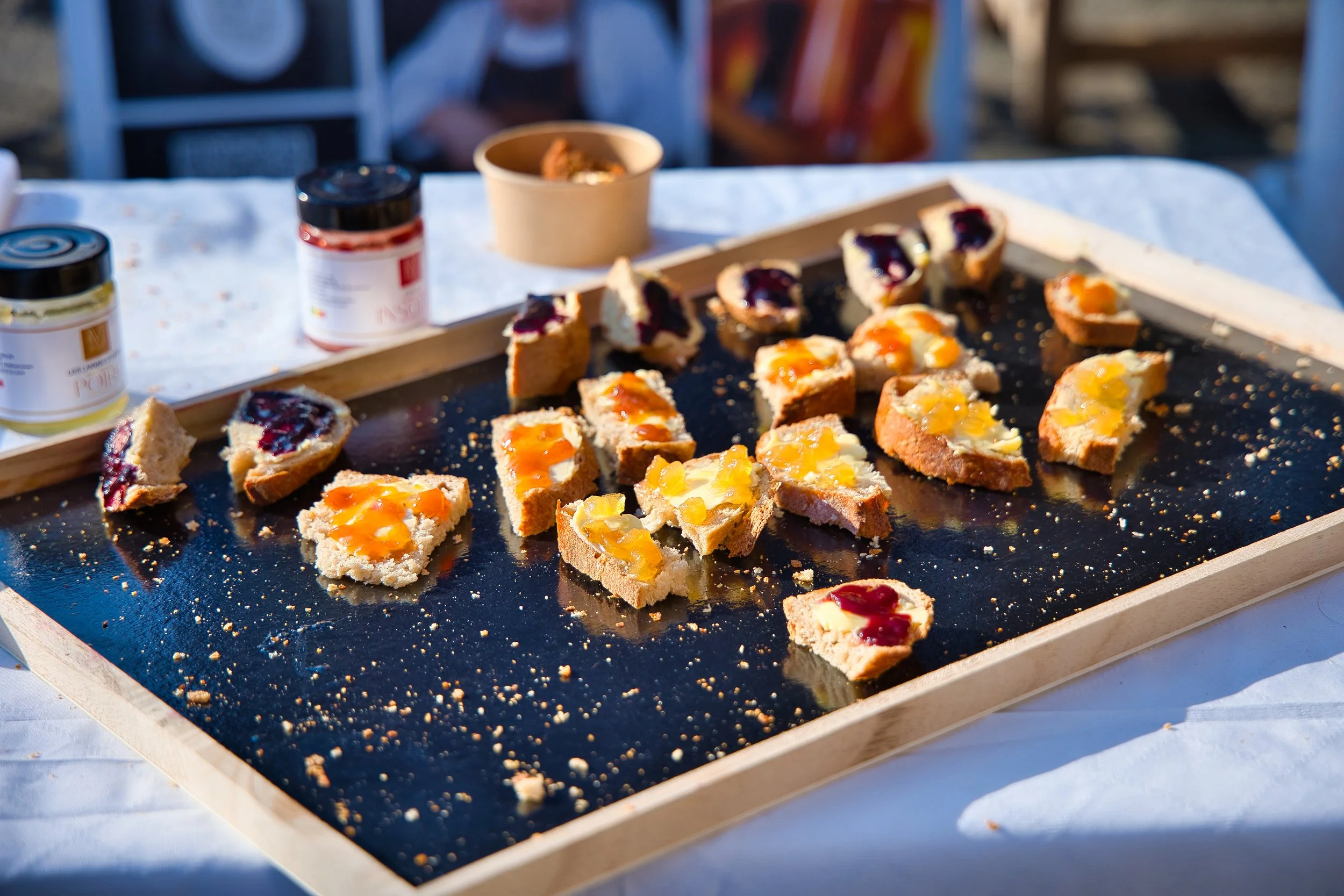 Petit toast avec confiture dans un plat en bois, sur une table blanche, avec des pots de confiture en arrière-plan.