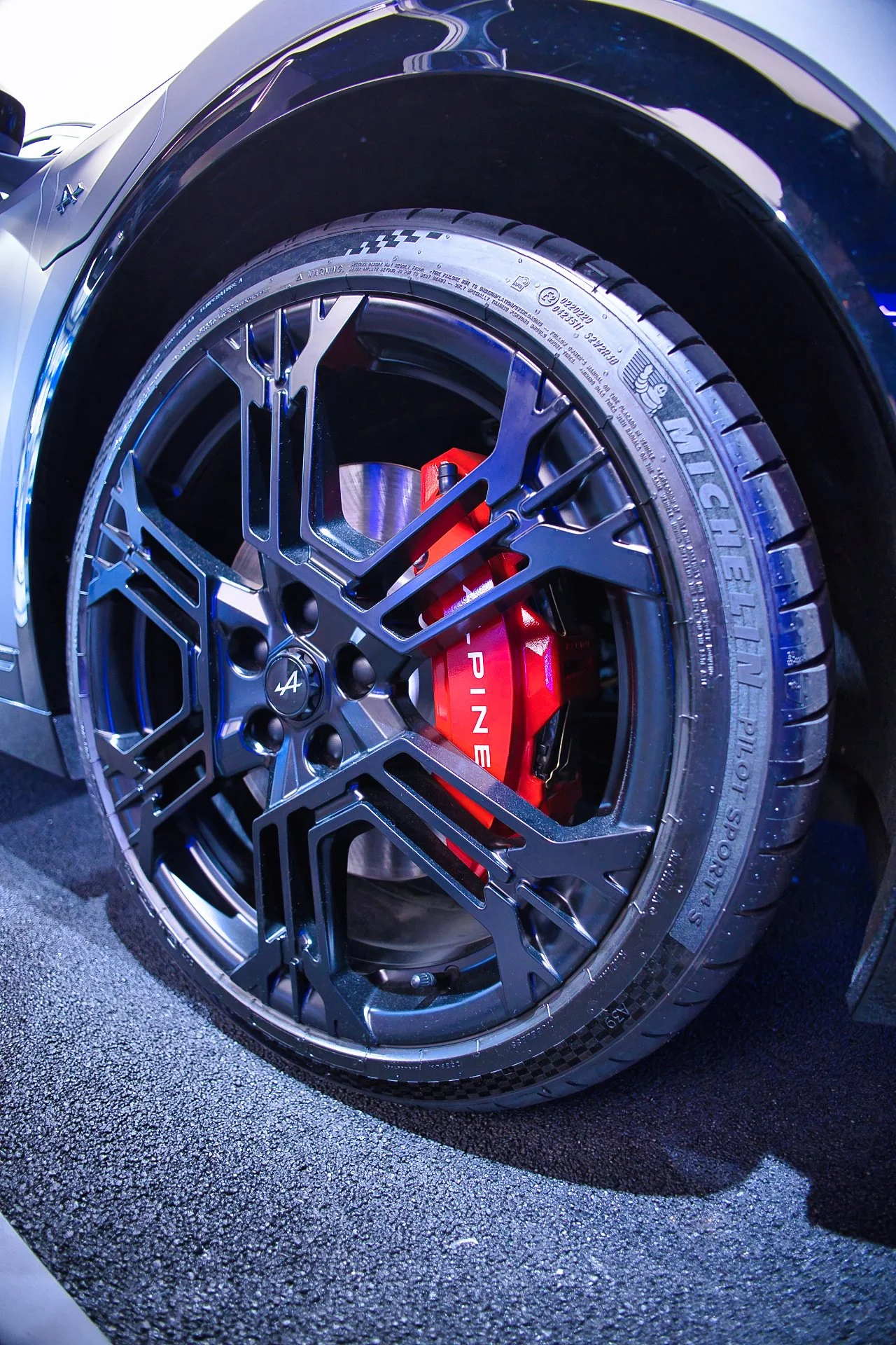 Un pneu de voiture Michelin monté sur une roue en alliage noir avec une calandre de frein rouge, affichant le logo 'Aline'. Alpine A390 à l'atelier Alpine Paris 