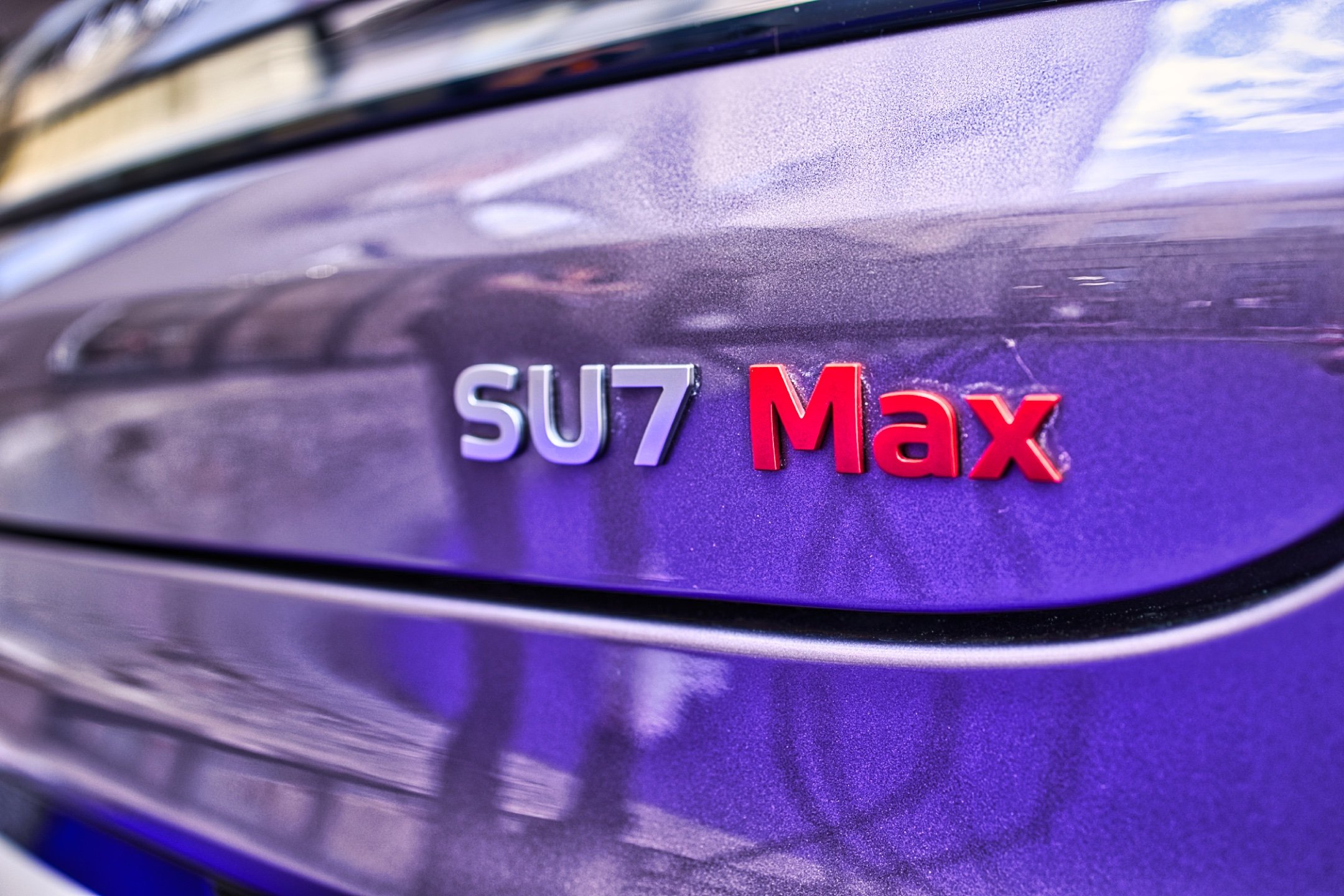 Gros plan sur le badge du modèle SU7 Max d'une voiture, avec un fond violet et un logo en relief rouge et blanc. Xiaomi SU7 Max
