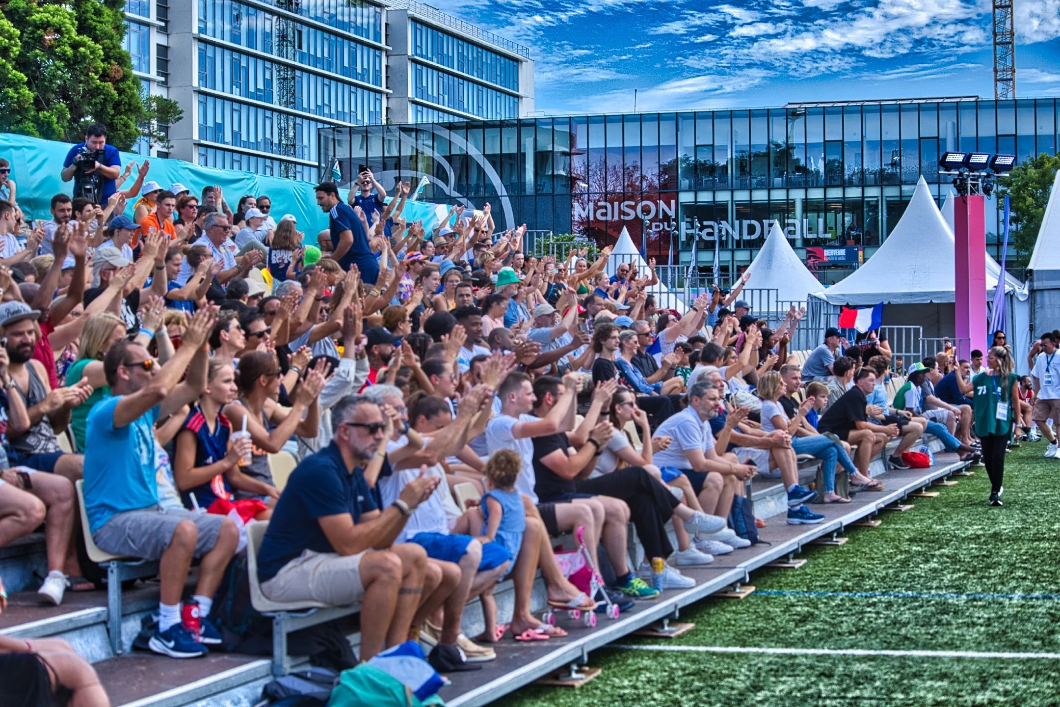 Spectateurs assis dans les gradins d'un match de Beach handball, certains applaudissant, sous un ciel bleu avec nuages, devant un bâtiment moderne en verre lors des Jeux de Paris 2024