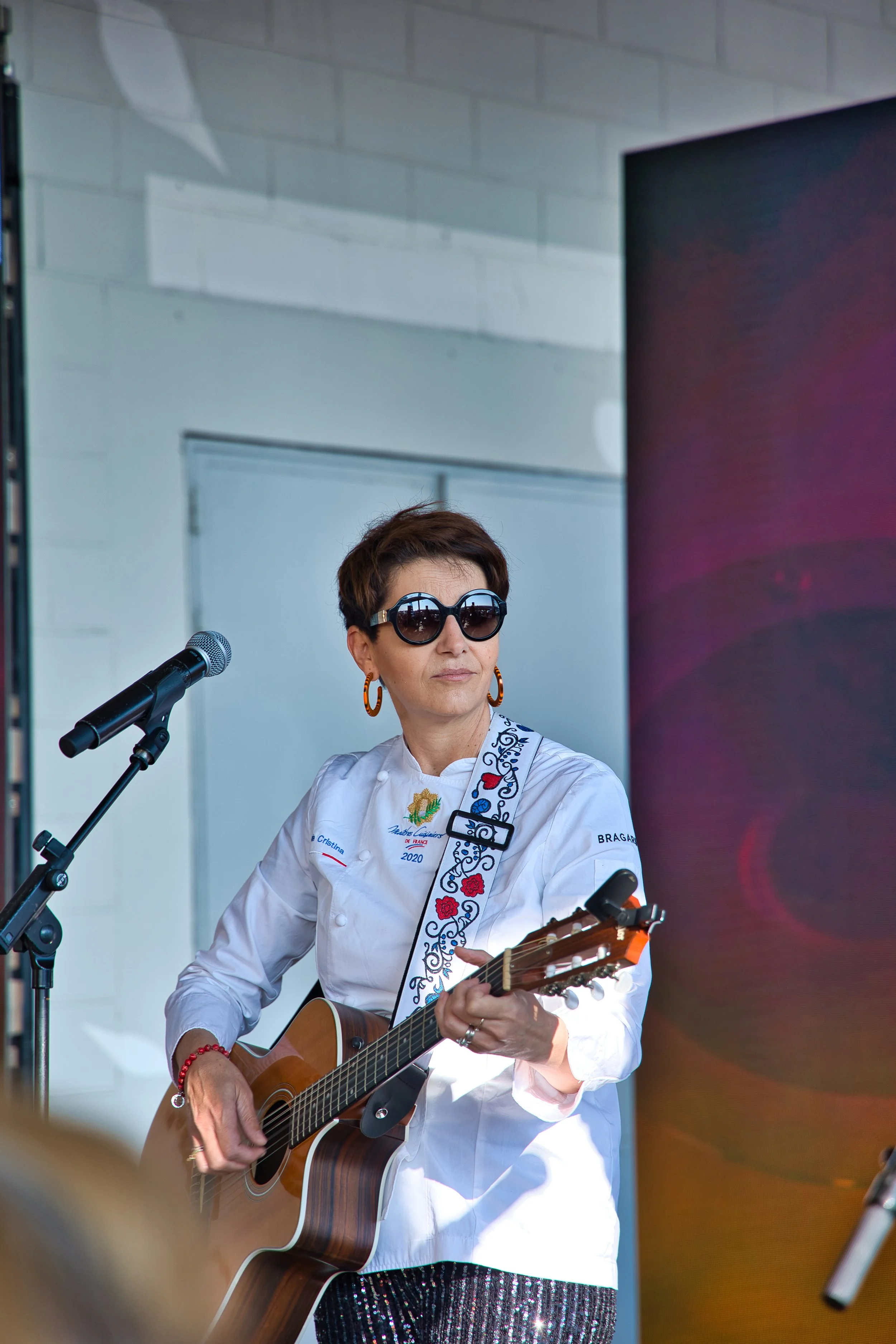 Une femme portant des lunettes de soleil, une chemise blanche et des boucles d'oreilles oranges joue de la guitare lors d'une performance.