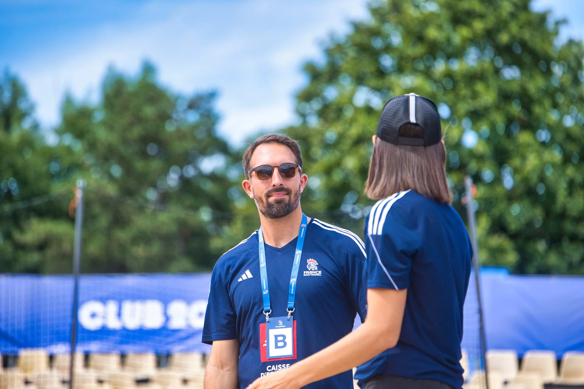 Deux personnes en tenue sportive discutent sur un terrain en plein air. L'homme porte des lunettes de soleil, un turban, un maillot de sport bleu et un badge. La femme, de dos, porte également un maillot bleu et un bonnet. JO Paris 2024