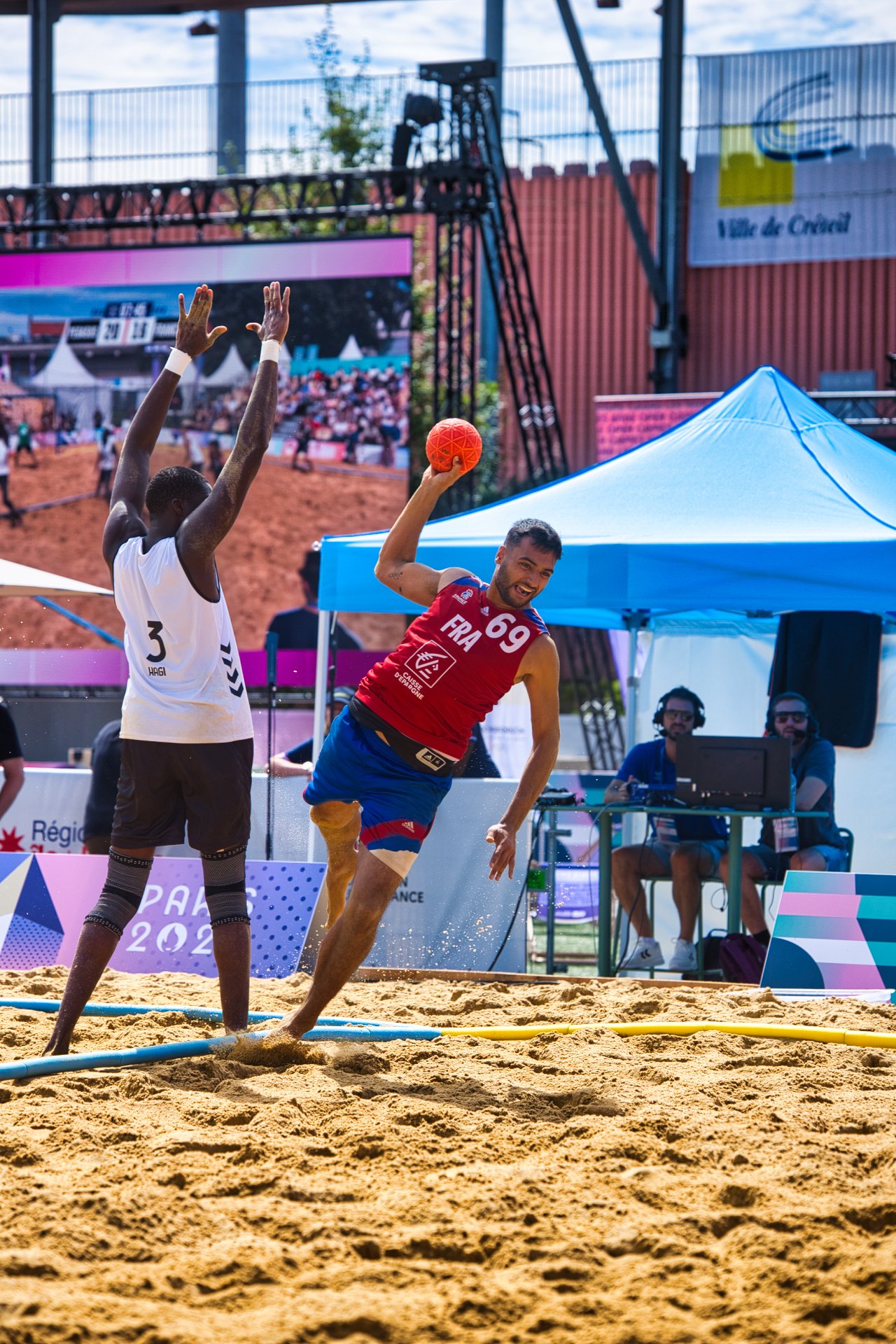 Un joueur de beach-volley en train de lancer le ballon en l'air, tandis qu'un adversaire tente de bloquer. Ils jouent sur un terrain de sable en plein air avec des spectateurs et des organisateurs en arrière-plan.