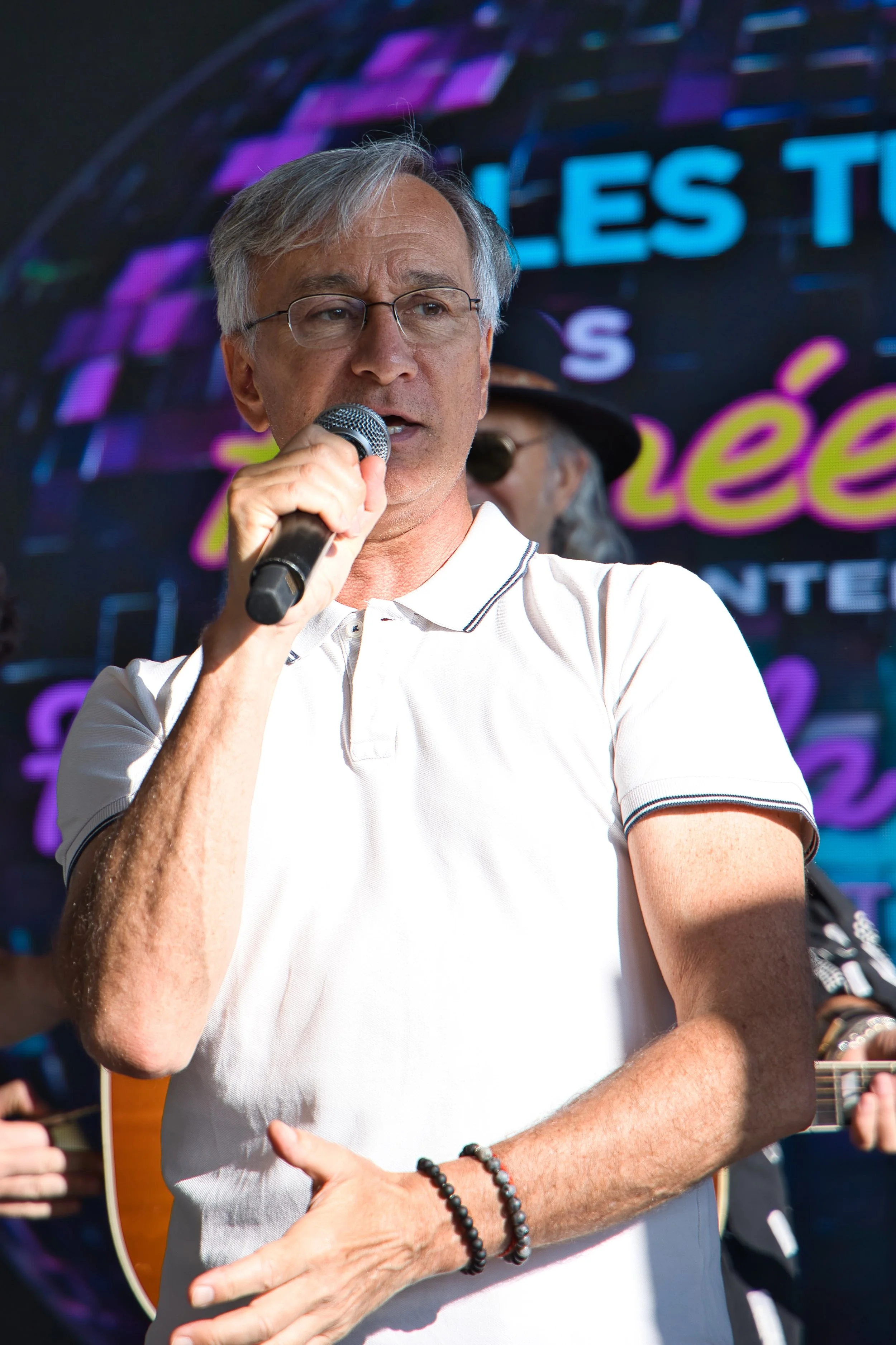 Un homme âgé avec des lunettes, portant un polo blanc, tenant un micro et parlant lors d'un événement en plein air, avec une guitare derrière lui et un écran lumineux en arrière-plan. fête de la musique au marché de Rungis