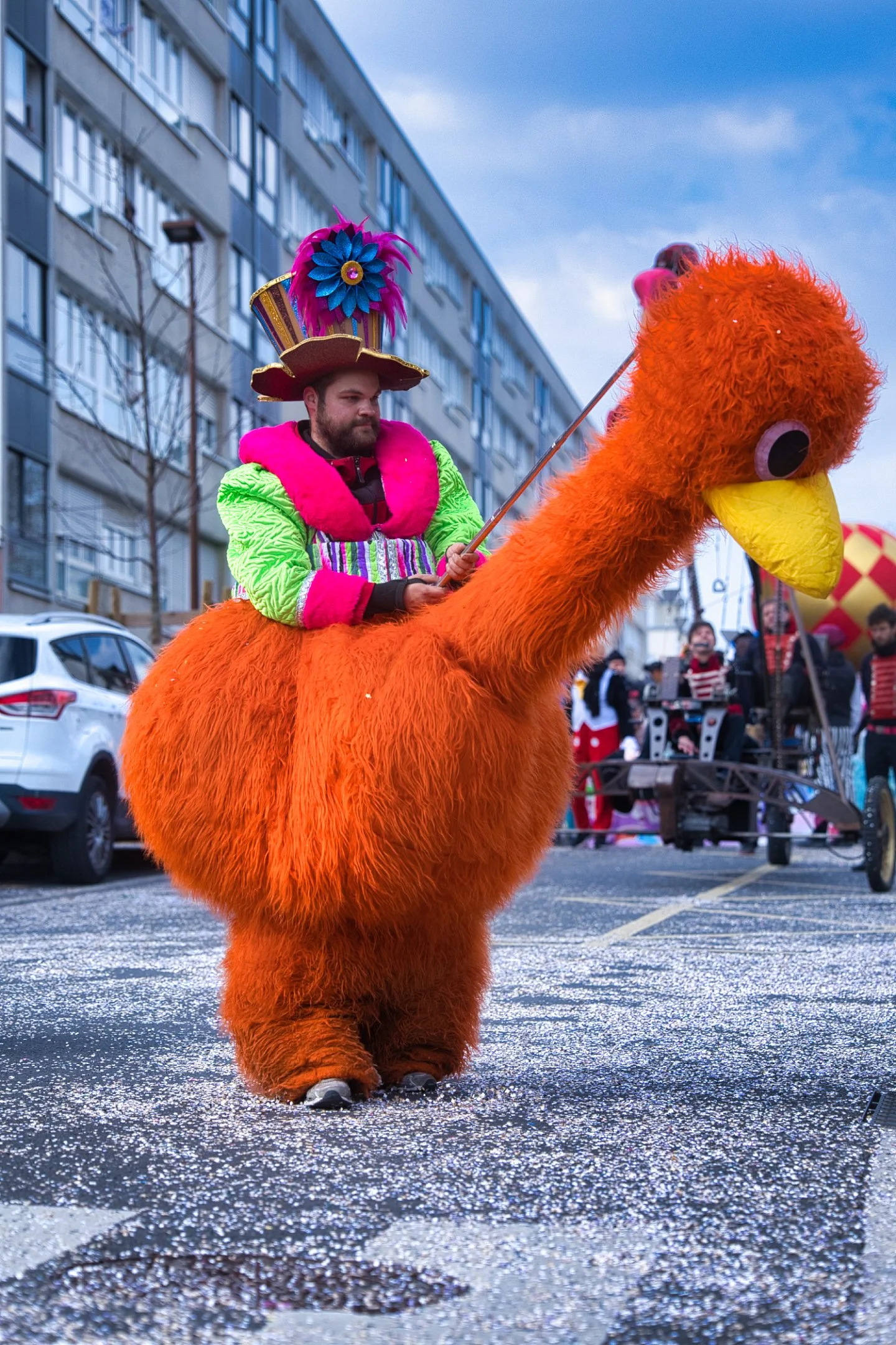 Une personne déguisée en personnage de canard géant orange, portant un costume coloré et un chapeau fantaisie, assise sur un animal en peluche géant. La scène semble être lors d'un carnaval dans une rue en ville.