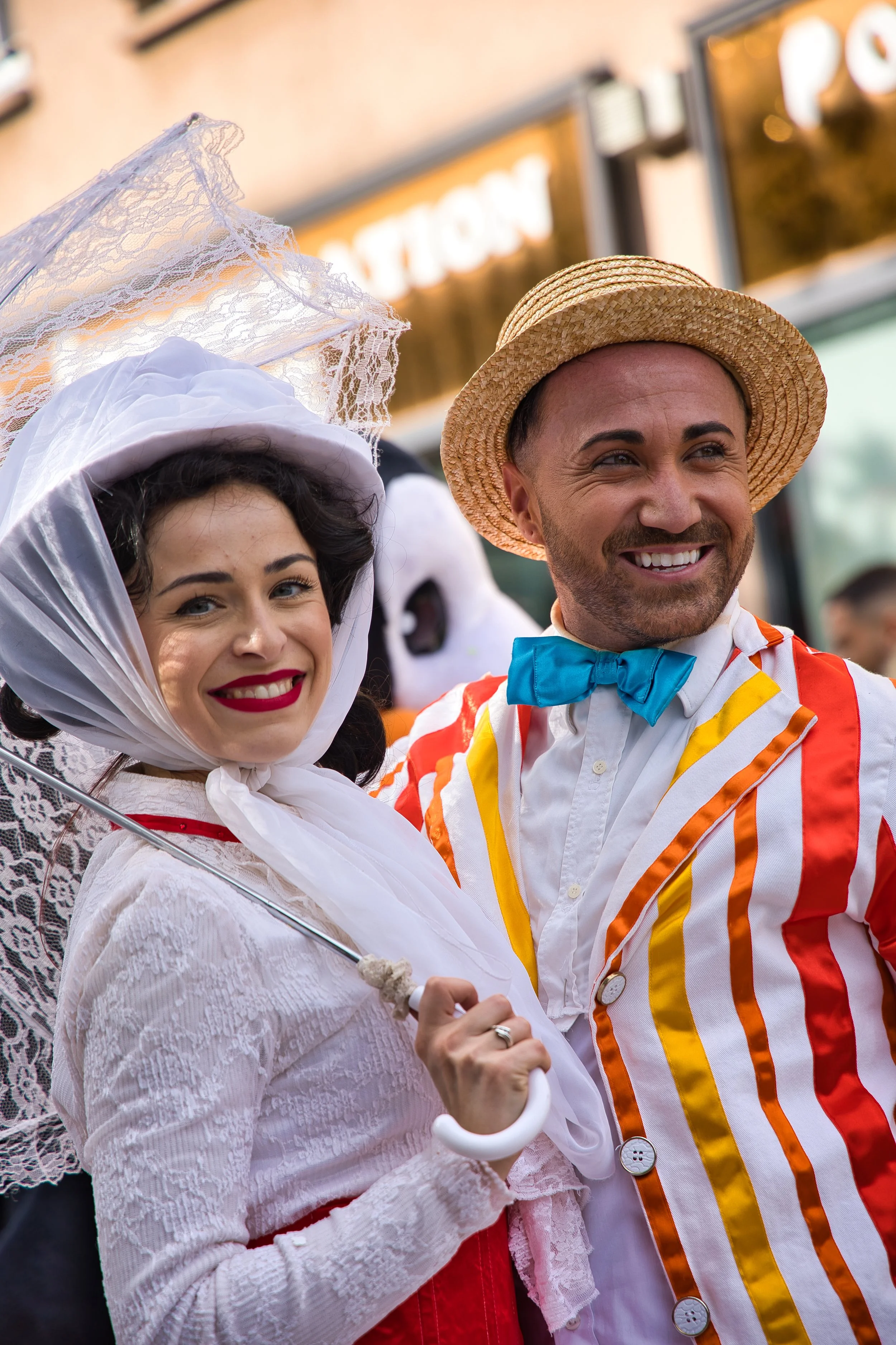 Deux personnes déguisées en costumes colorés et vintage, une femme avec un parapluie en dentelle blanche et une homme portant un chapeau de paille et un costume rayé orange, lors d'une fête en extérieur. carnaval thiais
