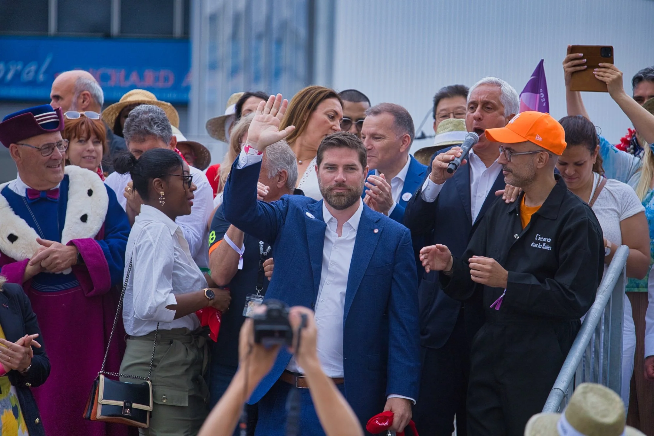Un groupe de personnes lors d'un rassemblement, un homme en costume bleu au centre faisant un signe de victoire, un autre avec un micro, un public diversifié avec des chapeaux et vêtements colorés, certains prennent des photos. marché de rungis