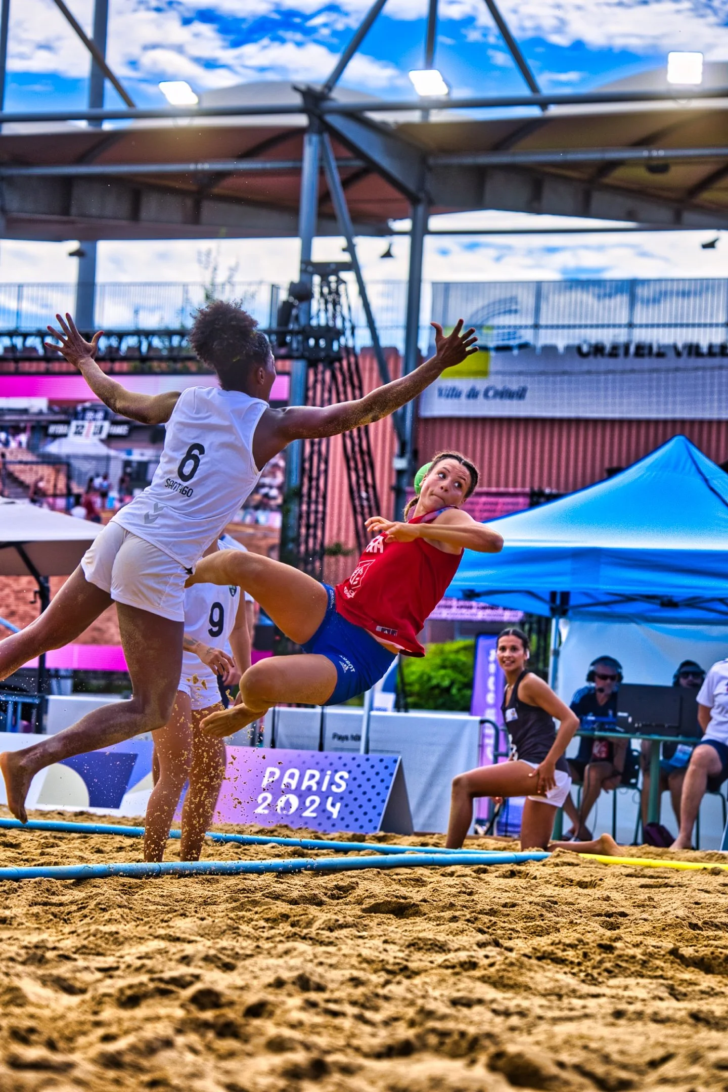 Deux joueuses de beach-volley en pleine action lors d'un match. La joueuse en blanc tente d'intercepter ou bloquer la joueuse en rouge qui s'apprête à frapper le ballon. L'événement se déroule en plein air avec un public en arrière-plan, sous un ciel