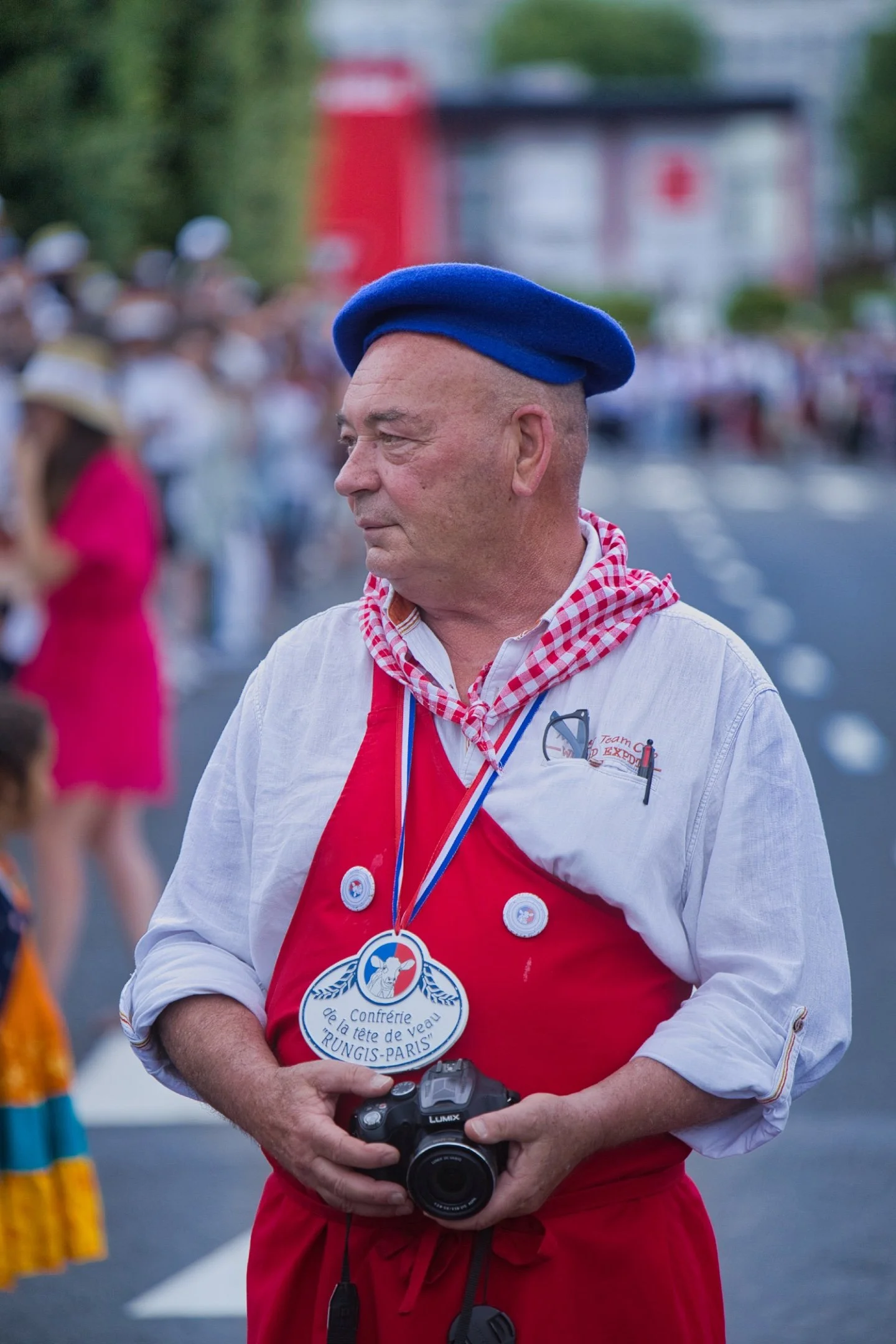 Un homme portant un béret bleu, un cordon de médaille, une chemise blanche et un tablier rouge, tenant un appareil photo, lors d'un événement en plein air avec un groupe de personnes en arrière-plan. cérémonie de la flamme olympique JO Paris 2024