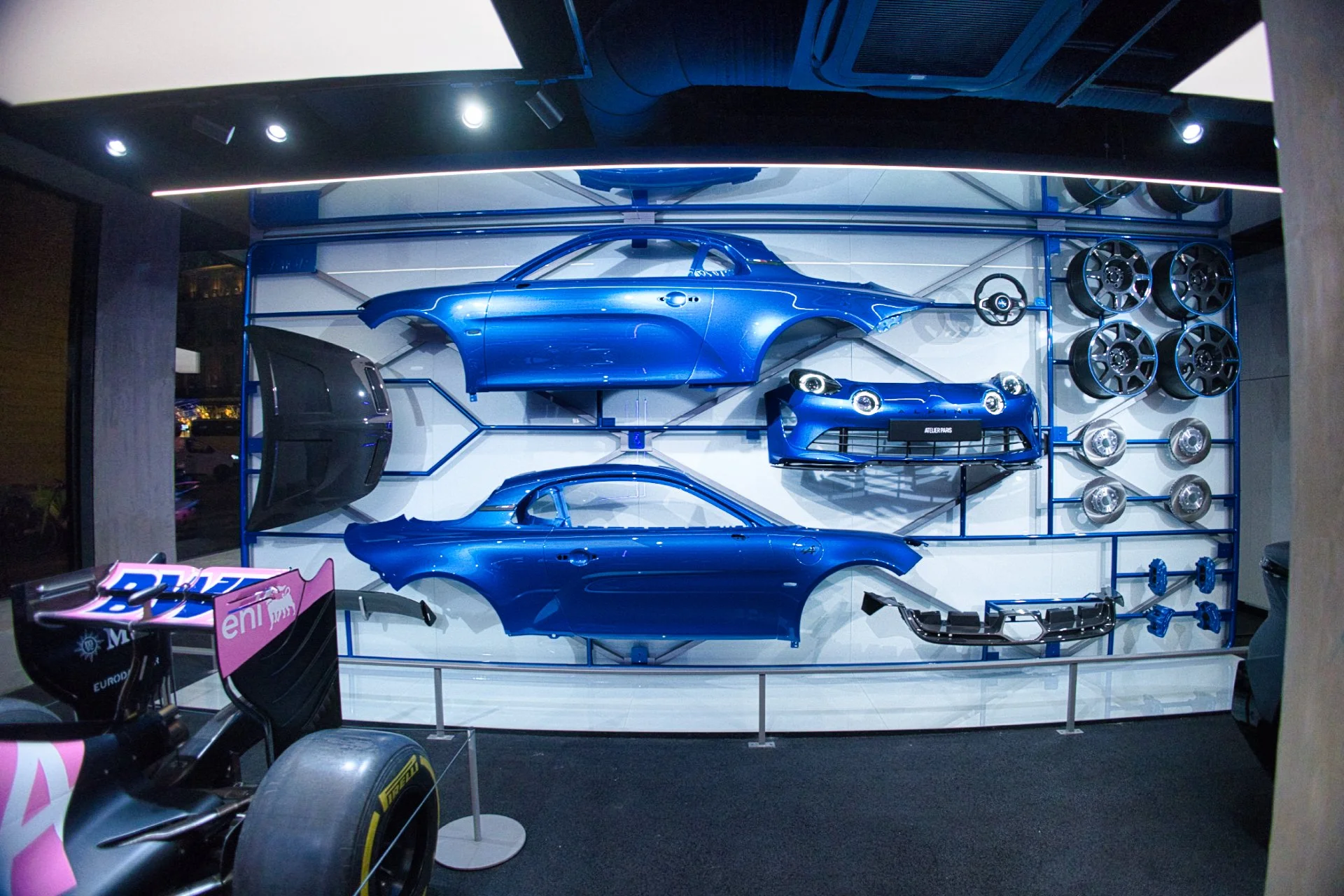 Exposition avec des pièces de l'Alpine A110, y compris des portières, capots, par teaux, roues et autres composants, assemblés sur un mur, avec une partie d'une voiture de course en premier plan à gauche à l'atelier Alpine à Paris