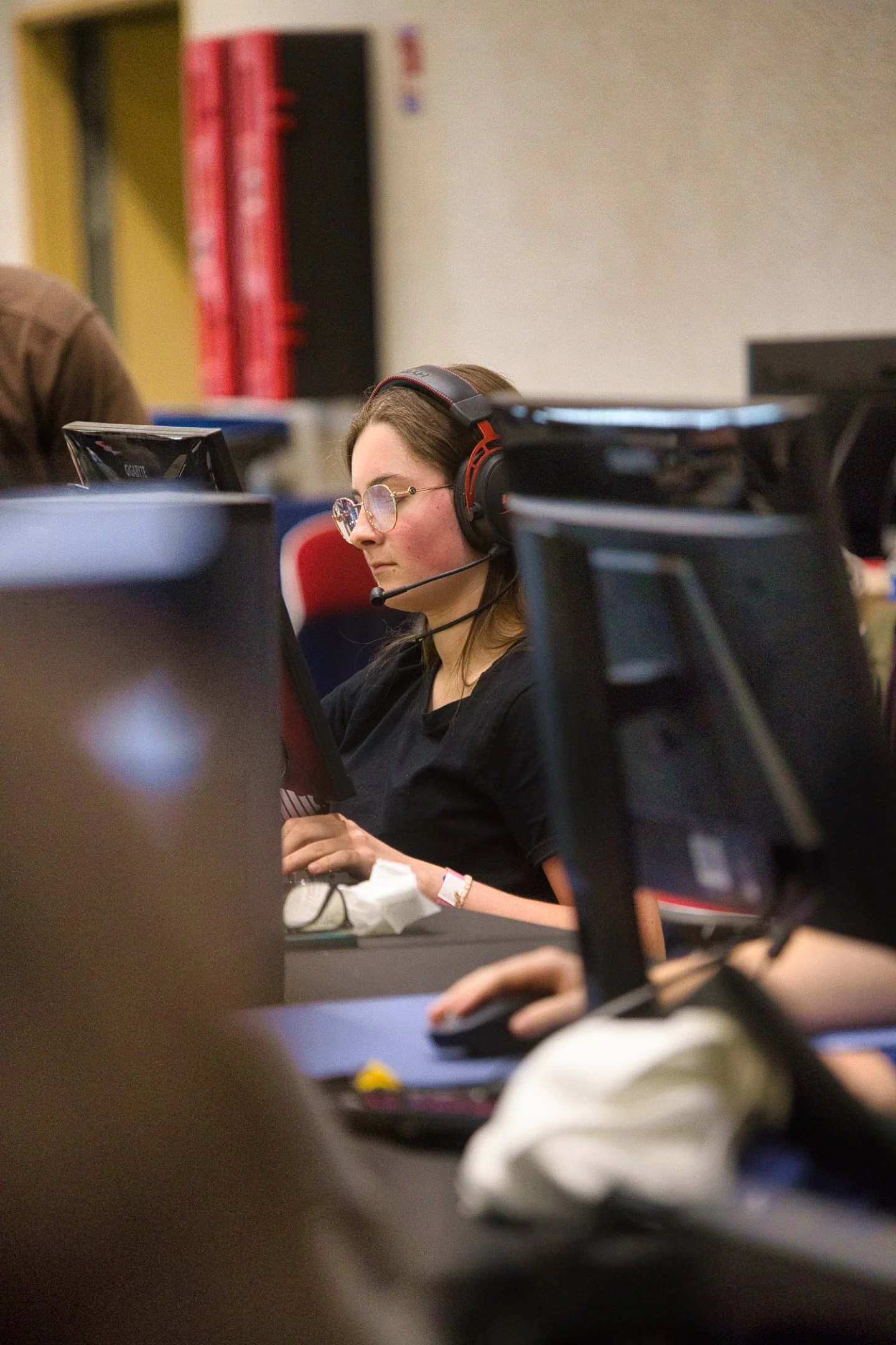 Une femme portant des lunettes et un casque avec microphone, concentrée devant un écran d'ordinateur lors de l'évènement Gaming Théodasium 2025 à Thiais