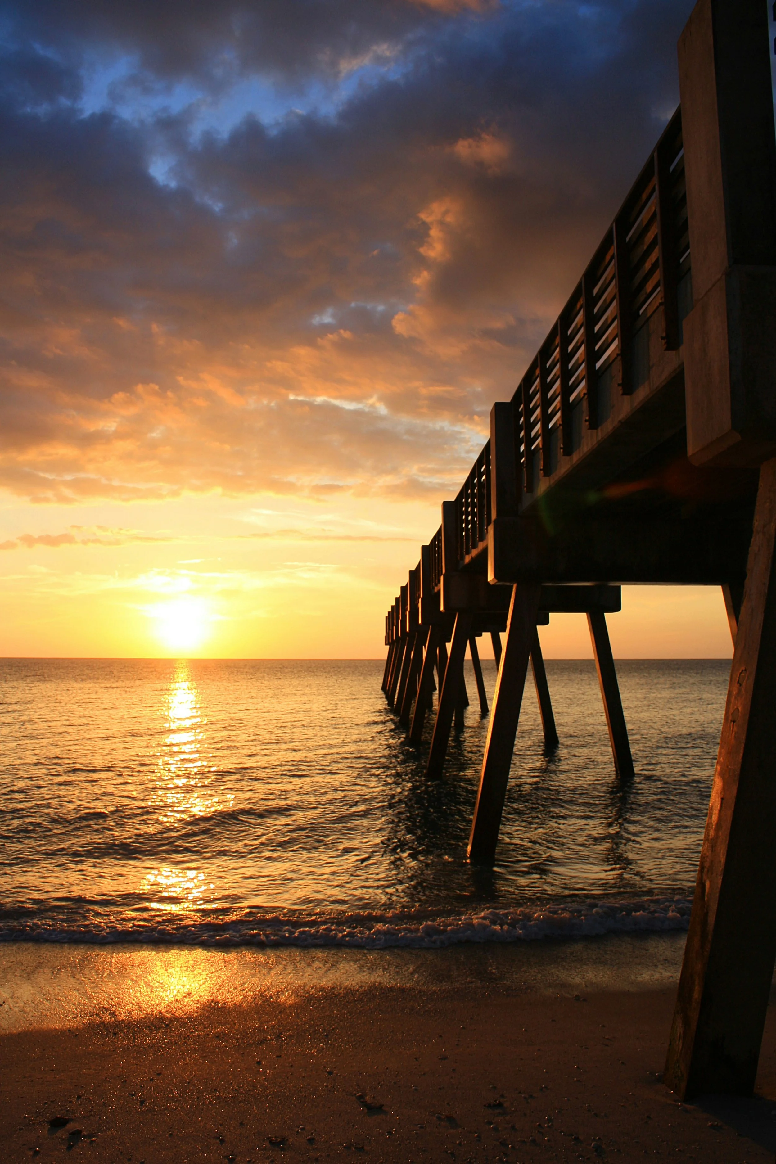 Sun setting over the ocean with a pier vero beach extending into the water, sky filled with clouds illuminated by the sunset, gentle waves lapping at the sandy shore.