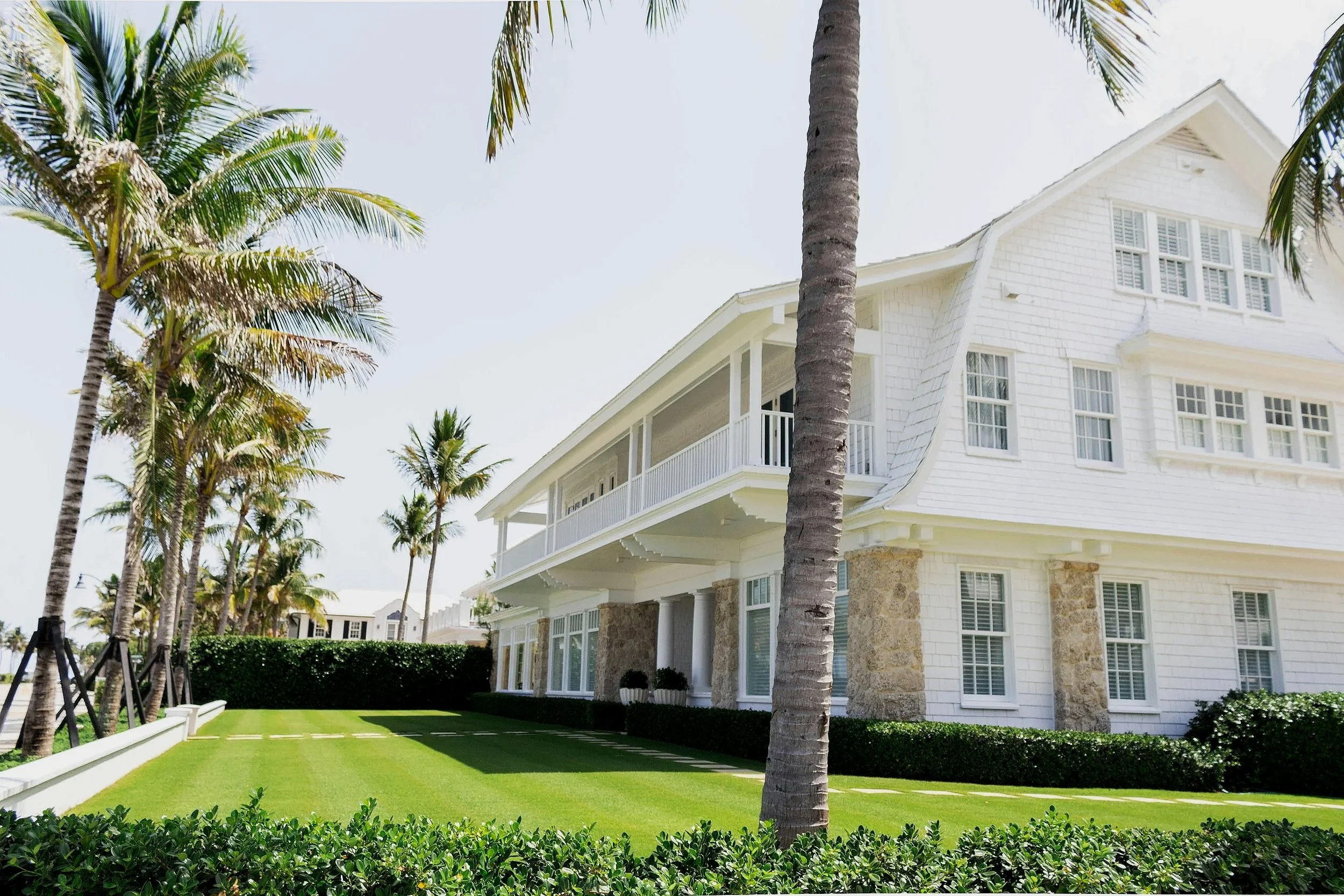 A white house with multiple windows and a balcony, surrounded by palm trees and a neatly trimmed lawn.