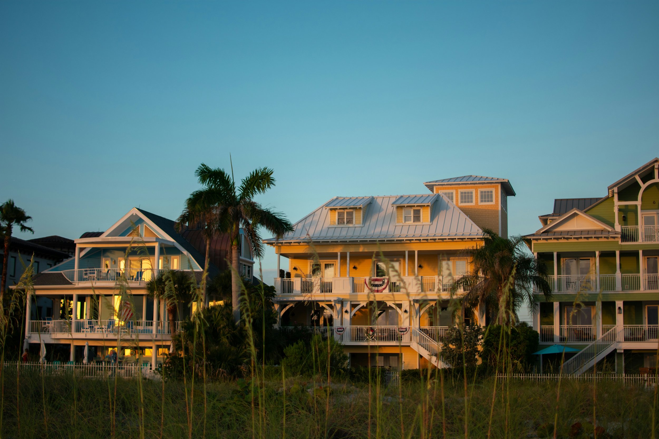 Three large beach houses with porches and stairs, palm trees, and tall grass in the foreground, illuminated by sunset light.