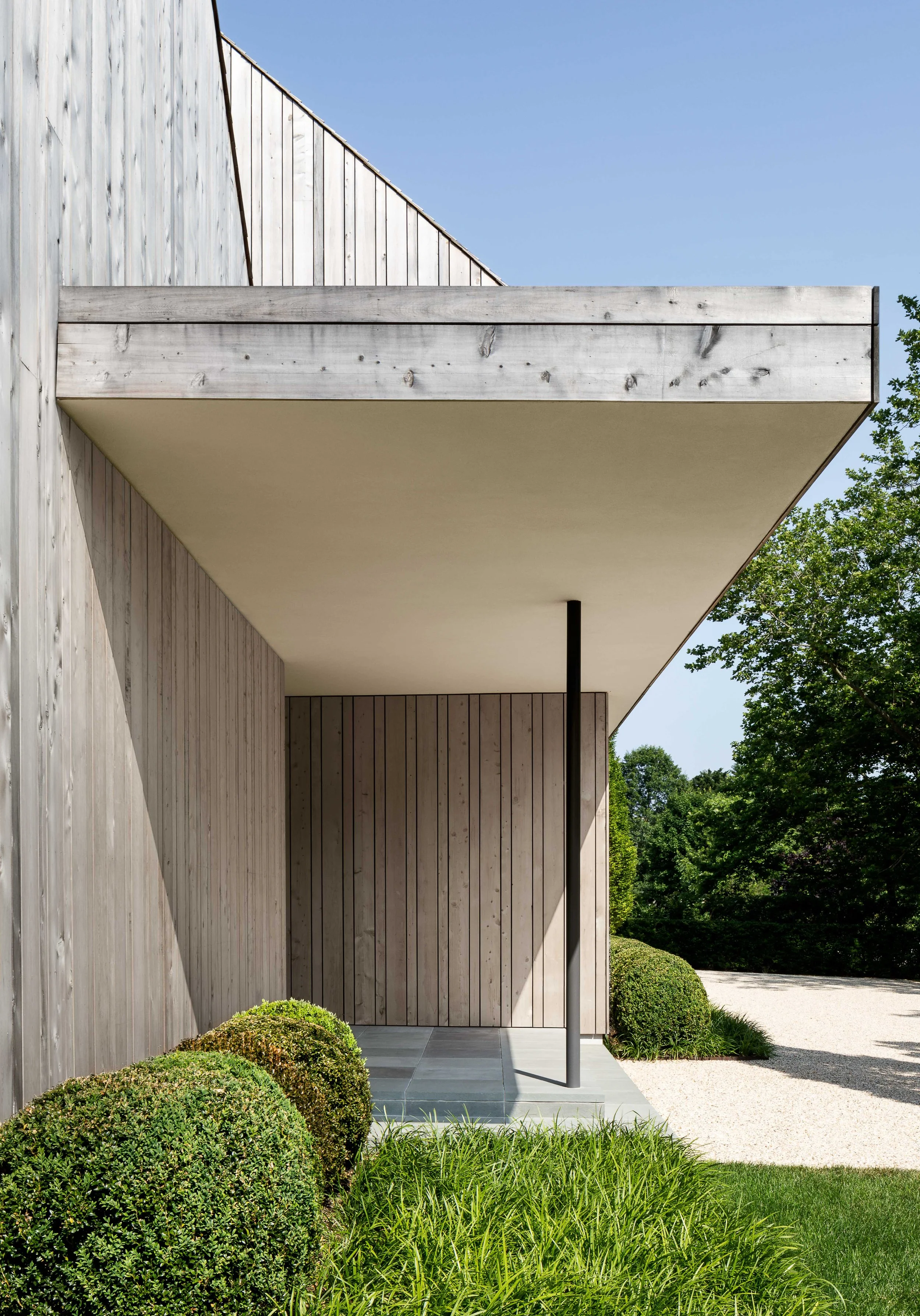 Modern house exterior with wooden paneling, a covered porch, and well-maintained green bushes and trees.