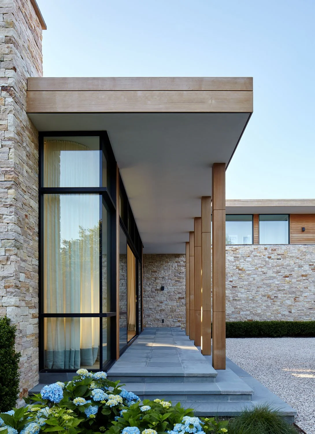 Modern house exterior with stone walls, large glass windows, and wooden accents, with blue hydrangeas in the foreground.