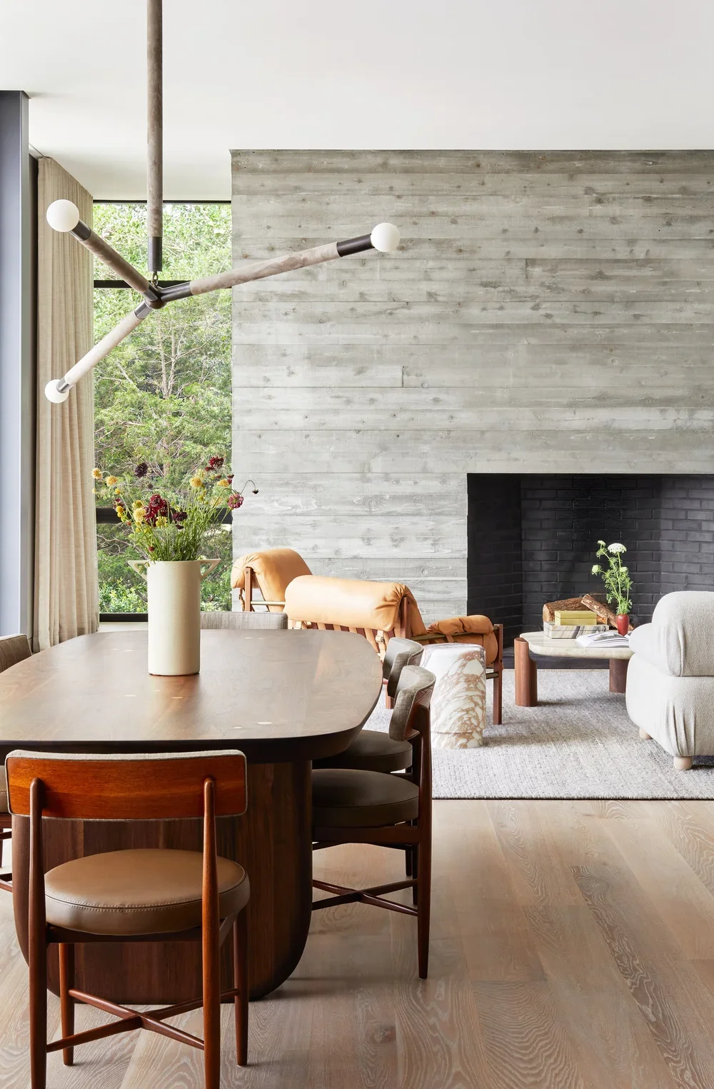 Modern living room with a wooden dining table, beige chairs, a large flower vase, a leather lounge chair, a gray sofa, a black fireplace, a coffee table, and large windows with greenery outside.