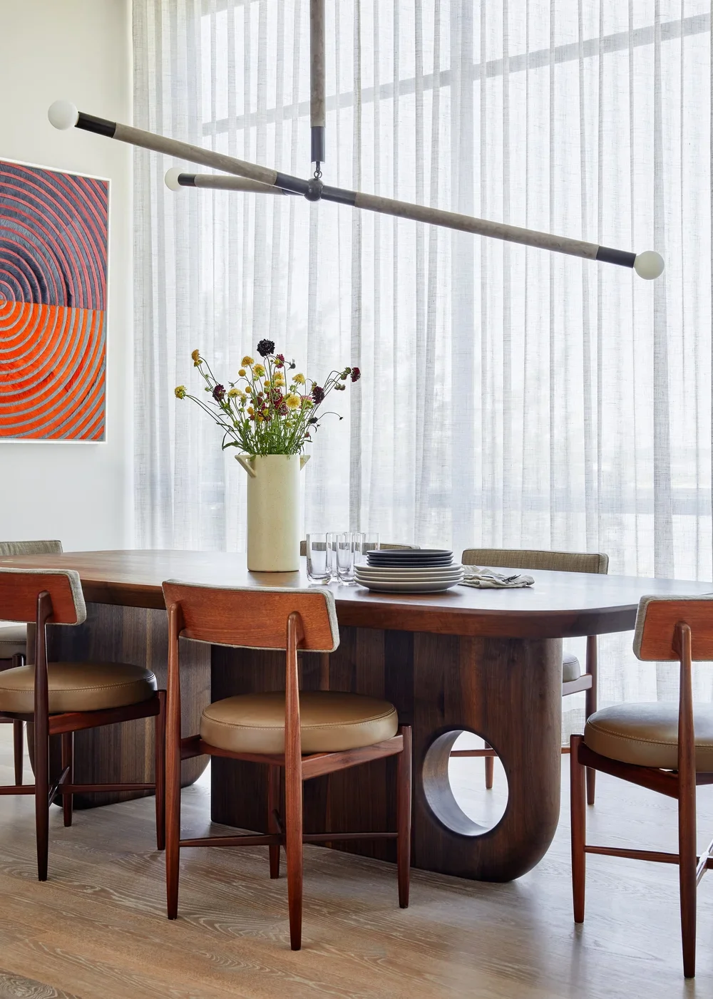 A modern dining room with a wooden table set with plates, glasses, and a white vase with flowers, surrounded by wooden chairs with cushioned seats, and a contemporary chandelier overhead, with sheer curtains and abstract art on the wall.