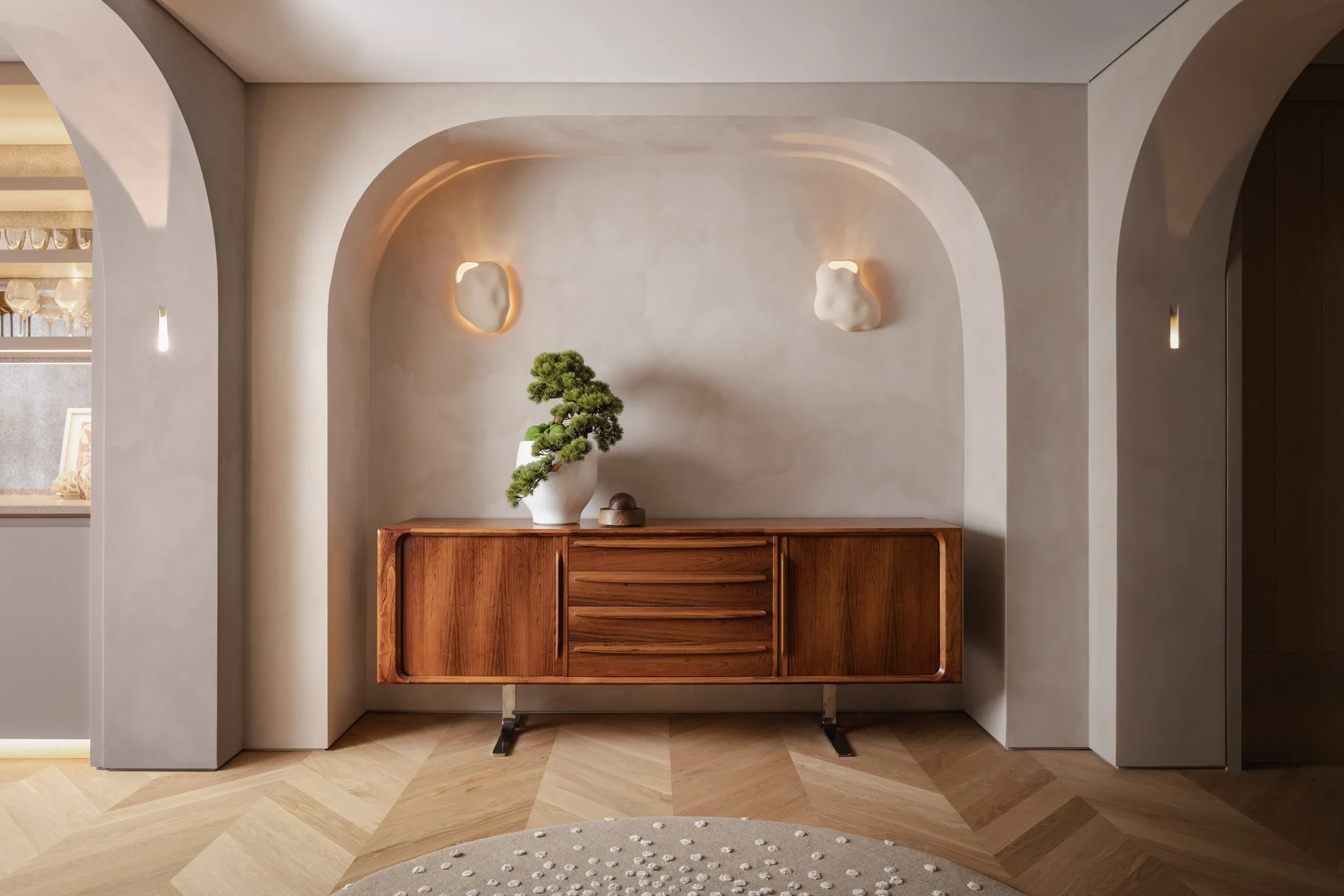 A wooden sideboard with a bonsai tree in a white pot, a small decorative object, and a rounded beige rug in front, set against a textured wall with modern wall sconces.