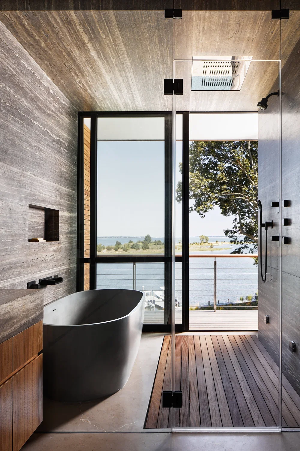 Modern bathroom with a freestanding bathtub, a large glass window with a view of water and trees, and a walk-in shower with a wooden floor and black fixtures.