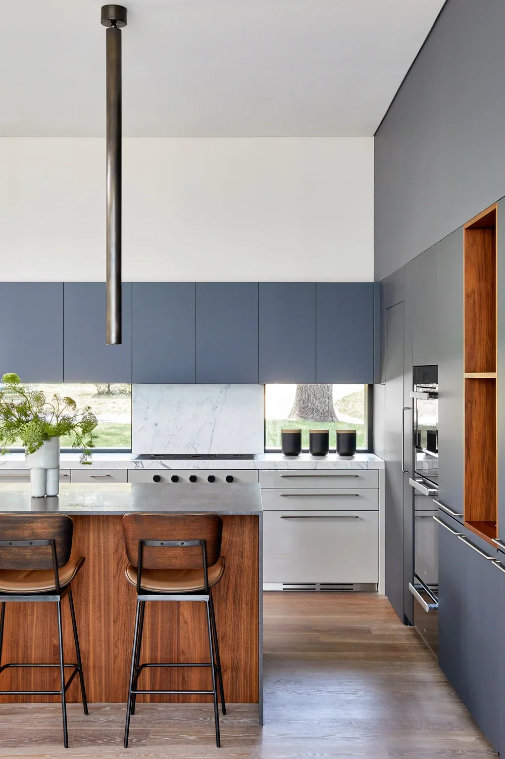 Modern kitchen with blue cabinets, white marble backsplash, wooden accents, and black decor, featuring a large window with outdoor views.