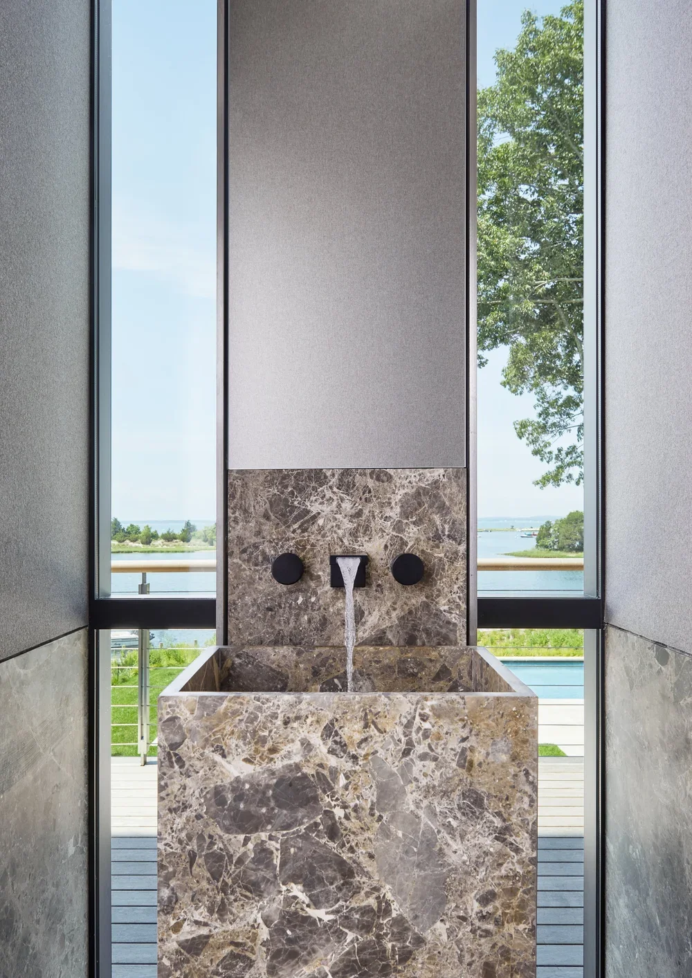 Interior of a modern building with a marble fountain on a balcony, large windows showing a scenic river and trees outside.