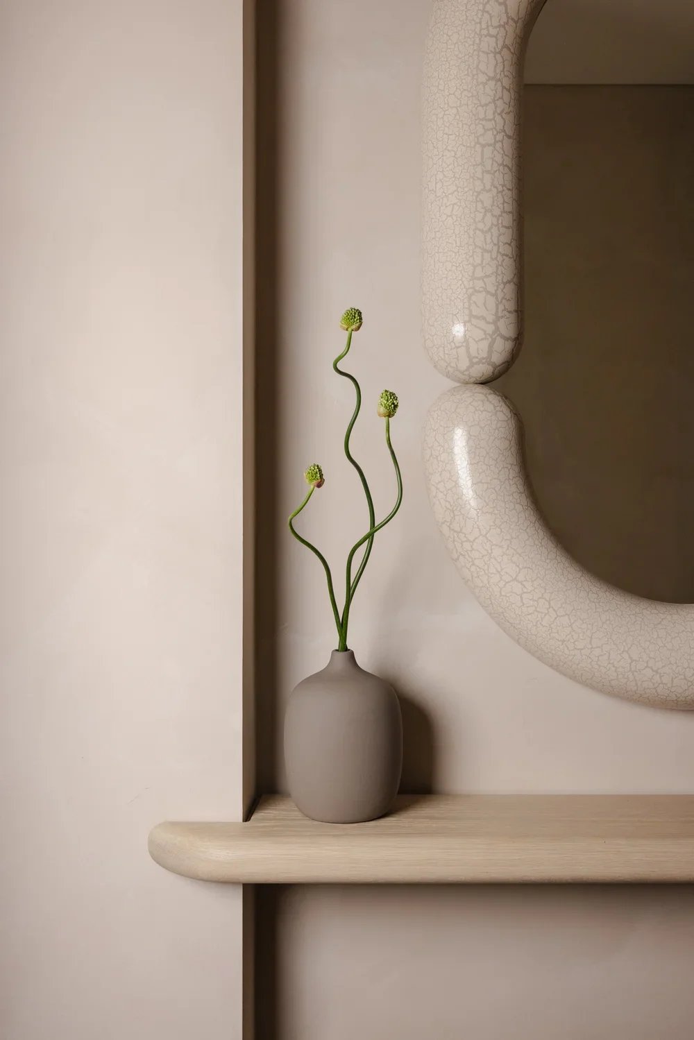 A beige vase with green, wavy stems and round flower buds on a light-colored shelf, next to a cracked cream-colored ceramic object and a rectangular mirror.