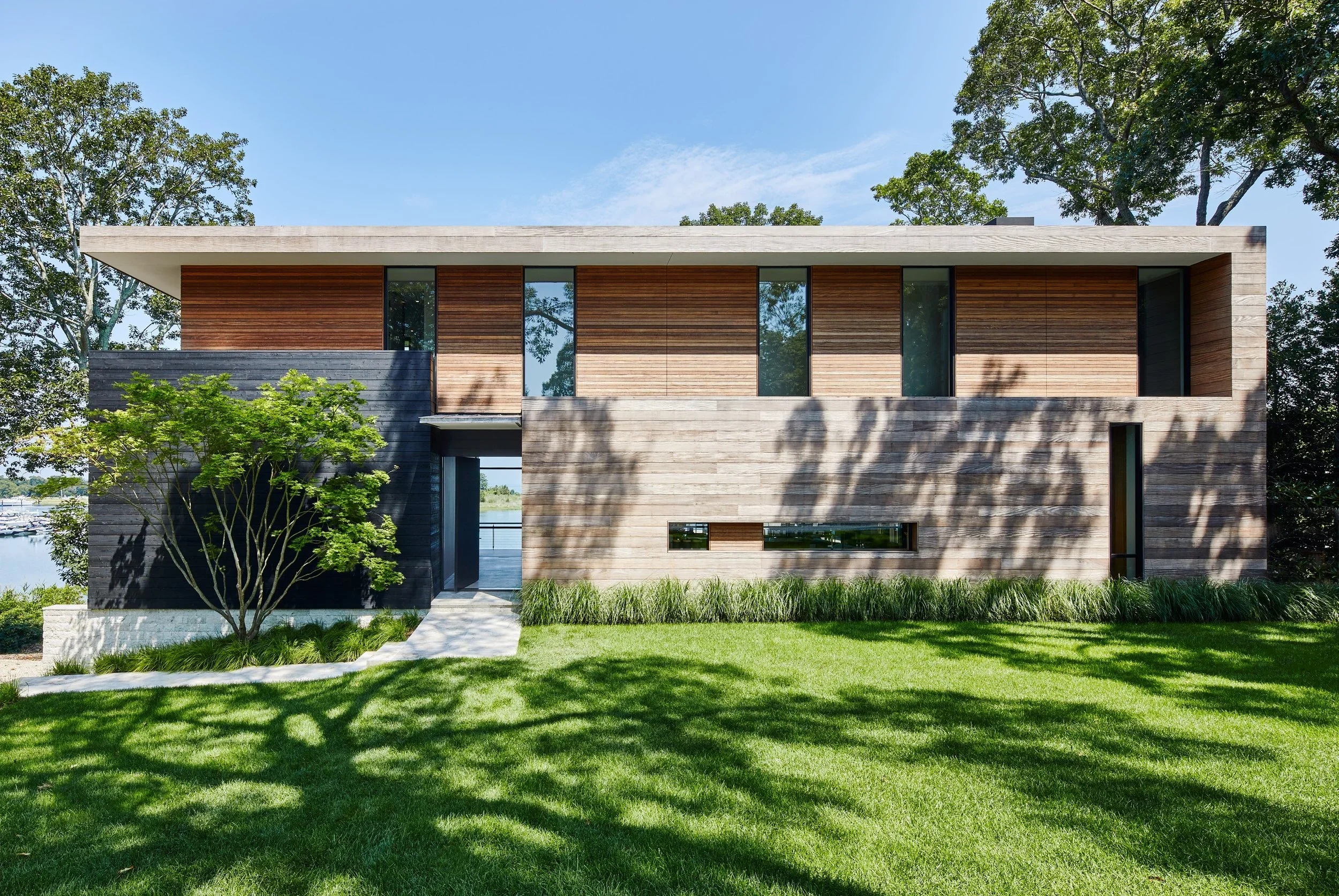 Modern two-story house with wood and concrete exterior, large windows, green lawn, and trees under a blue sky.