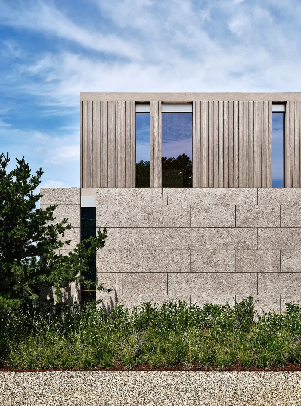 Modern building facade with wooden and stone elements, large reflective windows, surrounded by greenery and a blue sky with clouds.