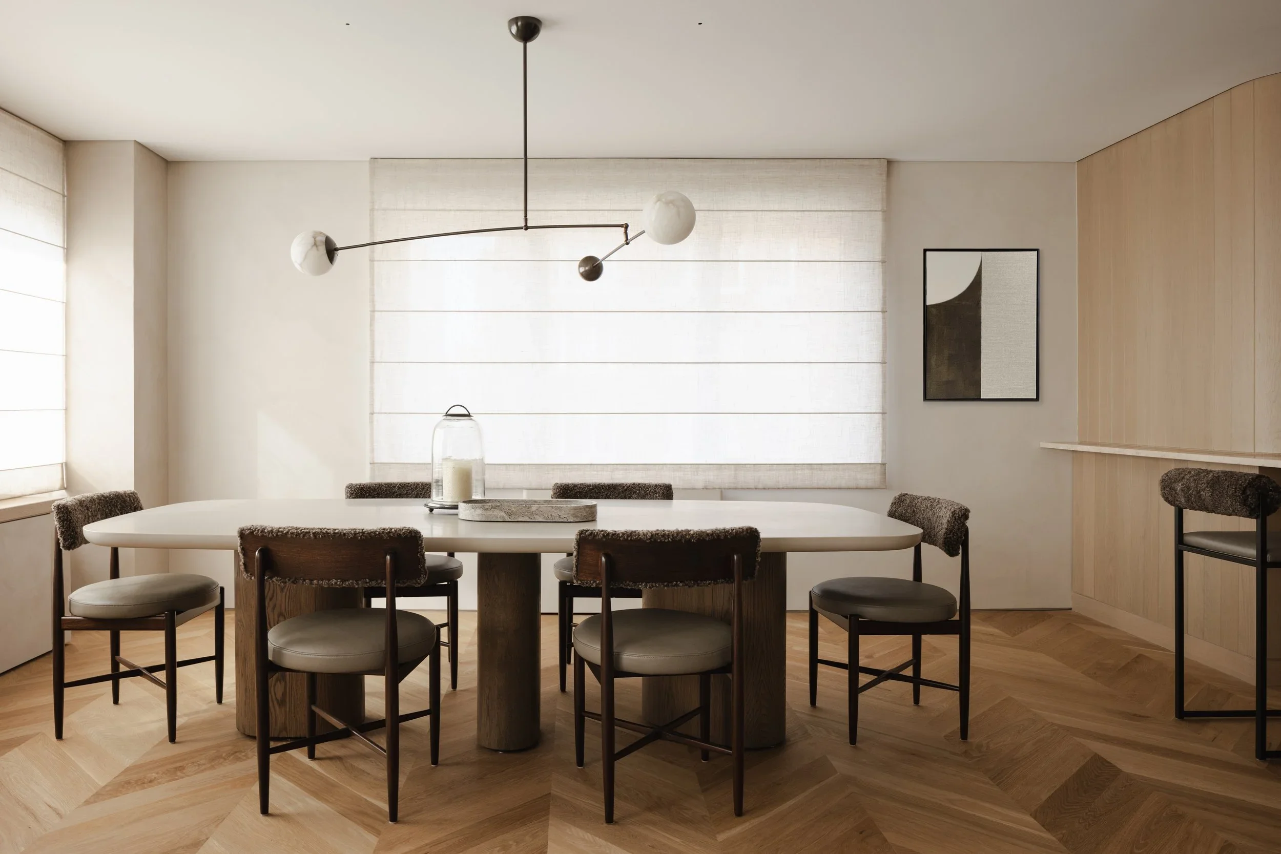 Modern dining room with a white oval table, six chairs with upholstered seats and brown furry backrests, and a contemporary chandelier overhead. The room has natural light from windows with white blinds, wooden flooring, a framed abstract artwork on 