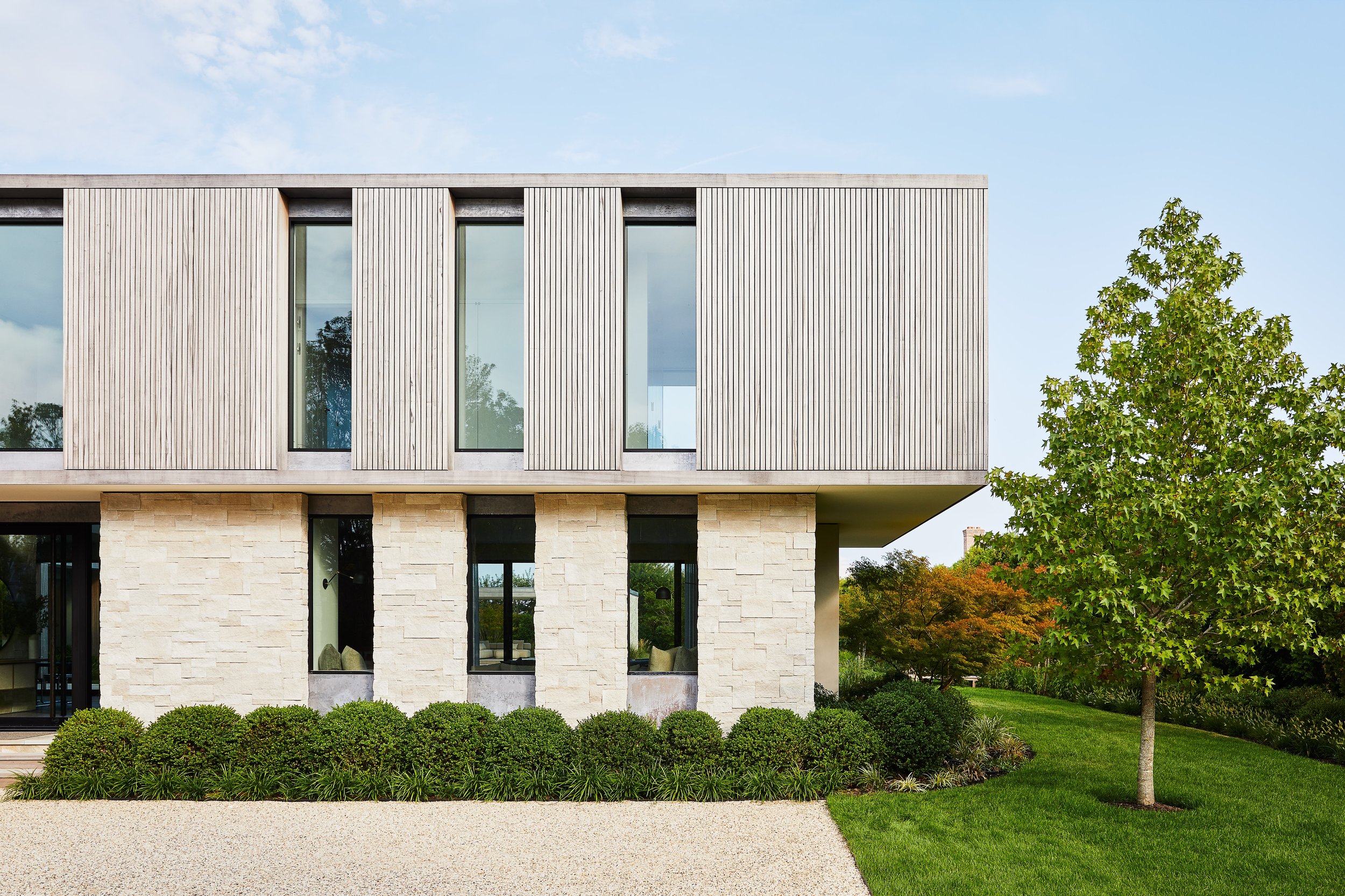Modern house with a beige stone lower level, large windows, and wooden vertical siding on the upper level, surrounded by green bushes, a grassy lawn, and a large tree to the right under a blue sky.