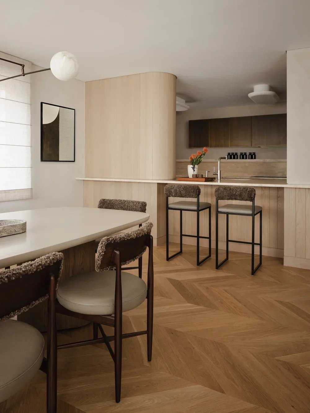 Modern kitchen and dining area with a white dining table, chairs, bar stools, and wooden cabinets, decorated with a flower vase and artwork.