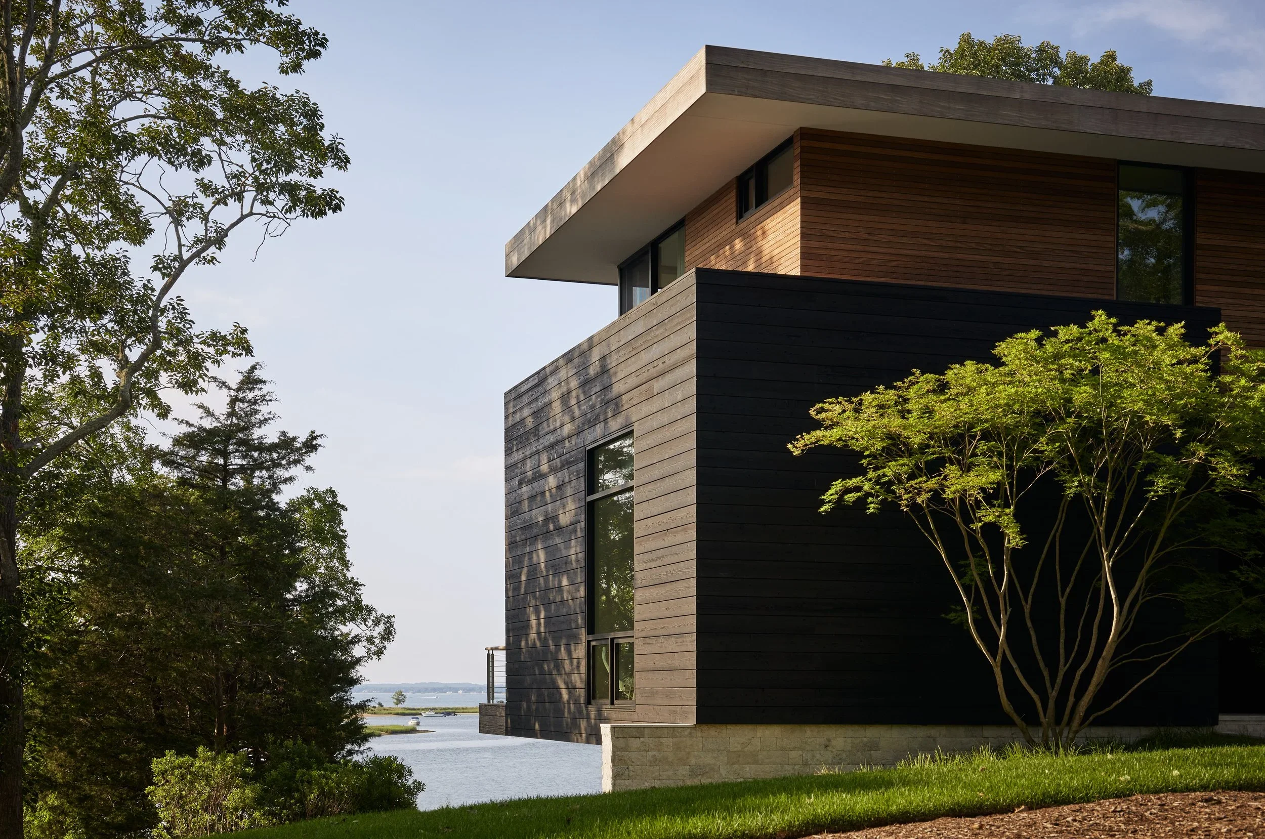 Modern house with black and wooden exterior, surrounded by trees, overlooking a body of water on a clear day.