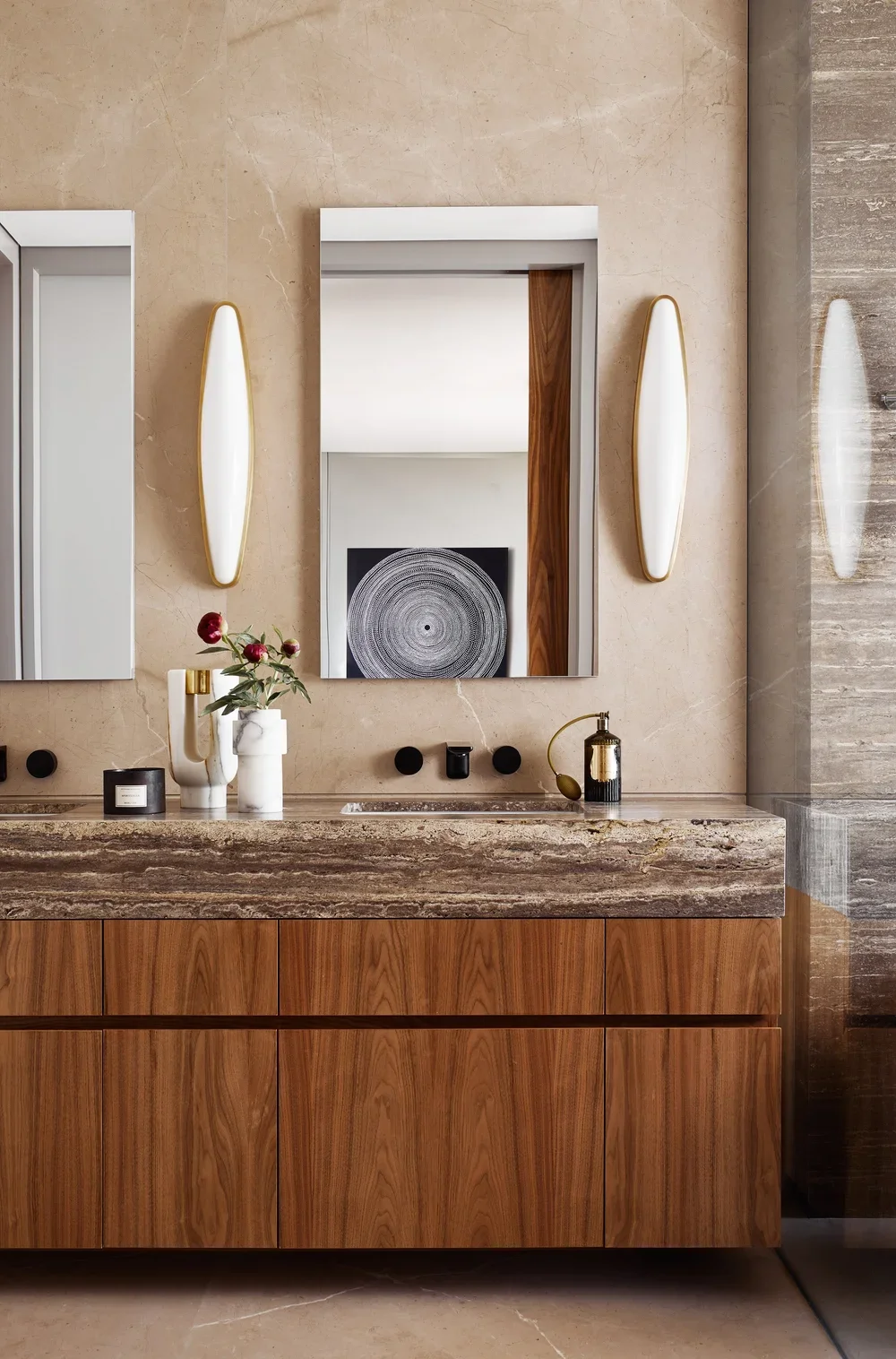 Modern bathroom vanity with a marble countertop, two mirrors, and decorative items including vases with flowers, a candle, and a soap dispenser, with lighting fixtures and textured wall surfaces.